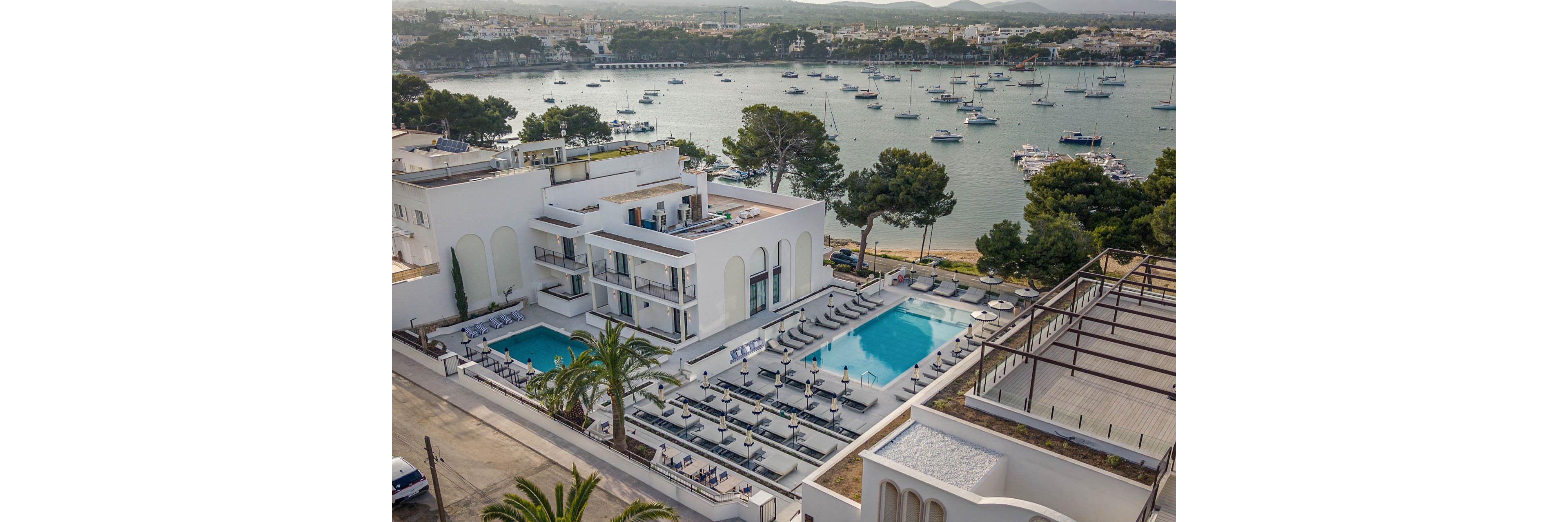 A modern white hotel complex with two outdoor swimming pools overlooks a calm waterfront marina. Numerous sun loungers and umbrellas surround the pools, creating a relaxed resort atmosphere. Sailboats and yachts are anchored in the bay, with a coastal town visible in the distance. Trees and palm trees frame the property, emphasizing a Mediterranean seaside vacation setting.