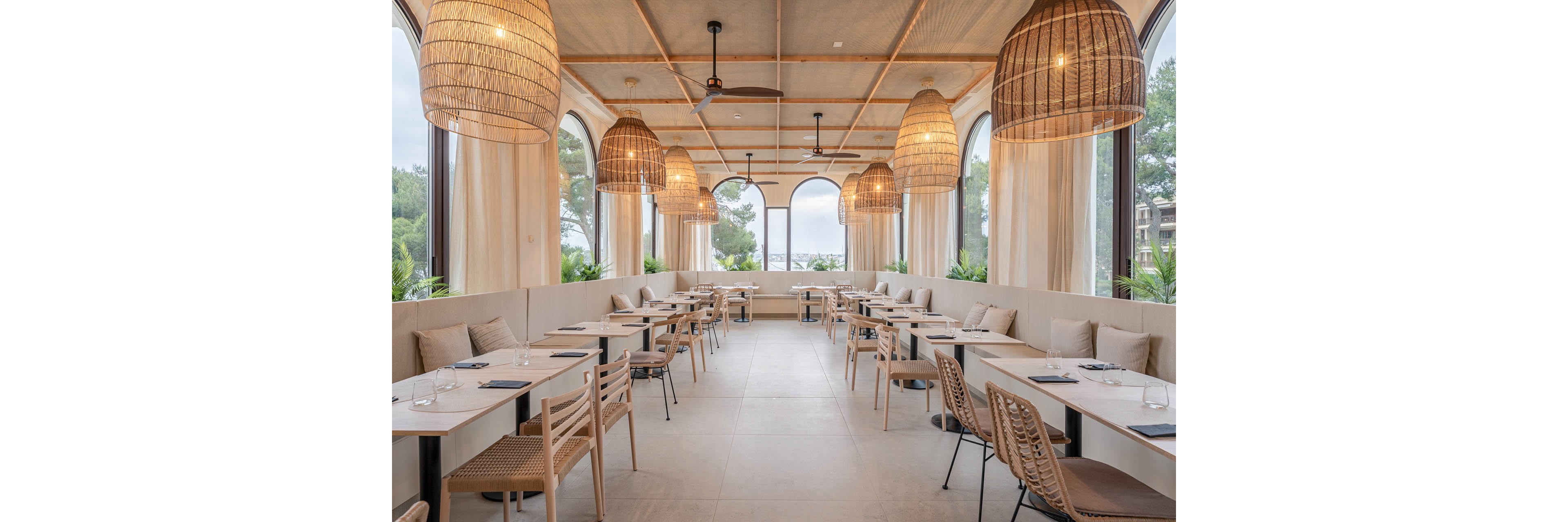 Contemporary restaurant interior featuring light wood tables and woven chairs is shown empty and ready for guests. Large arched windows line both sides of the space, revealing trees and a distant coastal view. Neutral beige tones, soft curtains, and cushioned bench seating create a calm, airy atmosphere. Oversized wicker pendant lamps and a ceiling fan add warmth and texture to the minimalist design.
