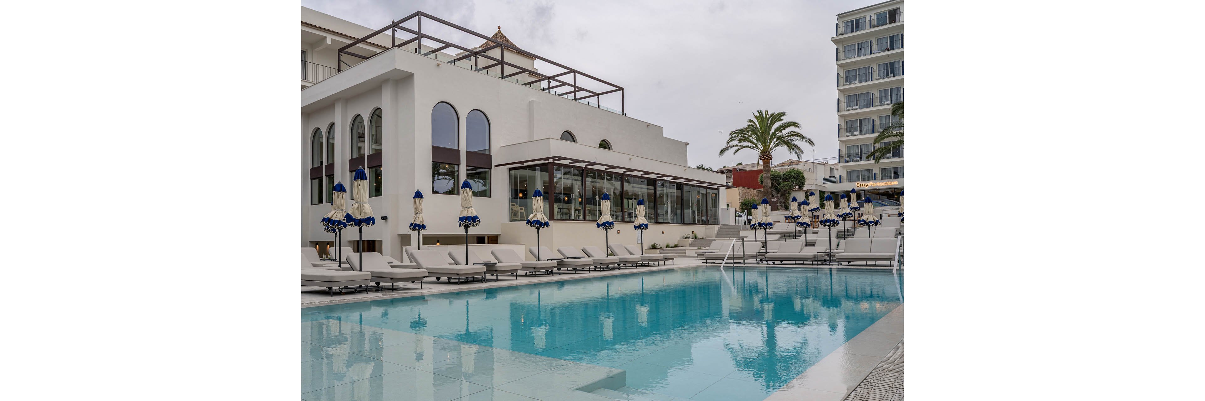 A large outdoor swimming pool sits in front of a modern white resort building with arched windows. Numerous sun loungers and closed umbrellas line the poolside terrace, suggesting a quiet off-peak moment. A tall palm tree and additional hotel structures are visible in the background, enhancing the vacation atmosphere. The overcast sky creates soft, diffused lighting over the calm blue water.