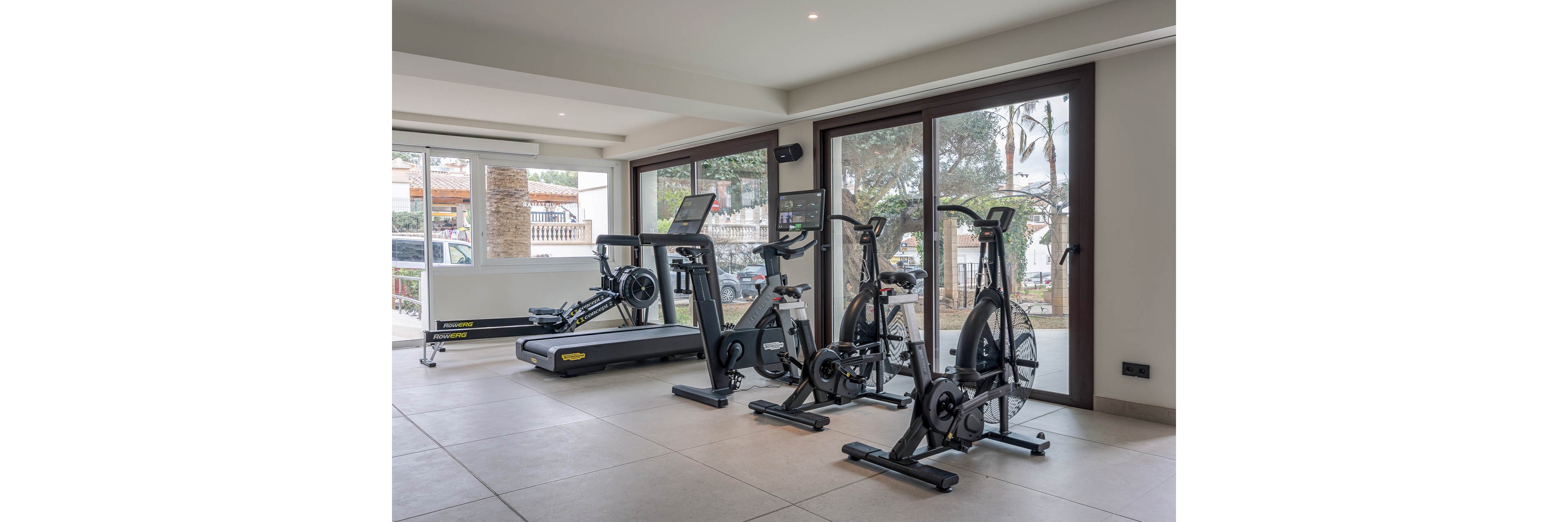 A bright home gym features several pieces of modern cardio equipment neatly arranged in front of large glass doors. The room includes a treadmill, rowing machine, stationary bike, and air bike, all facing inward for convenient use. Natural light streams through the windows, revealing trees and an outdoor area beyond. The clean, minimalist interior and neutral color palette create a calm, contemporary workout space.