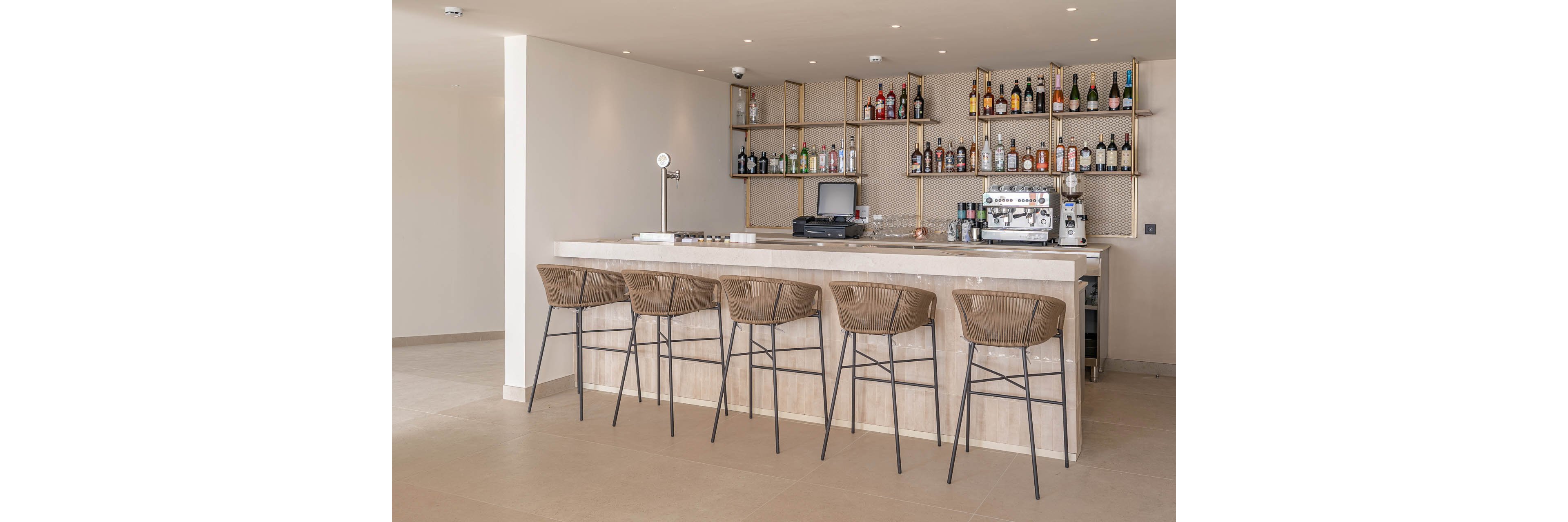 A contemporary bar counter stands in a bright, minimalist interior. Five high bar stools line the light wood counter, facing a neatly organized back bar with bottles and glassware. A beer tap, espresso machine, and other bar equipment are visible on the countertop. Neutral tones and clean lines create an airy, modern atmosphere suitable for a hotel or resort lounge.