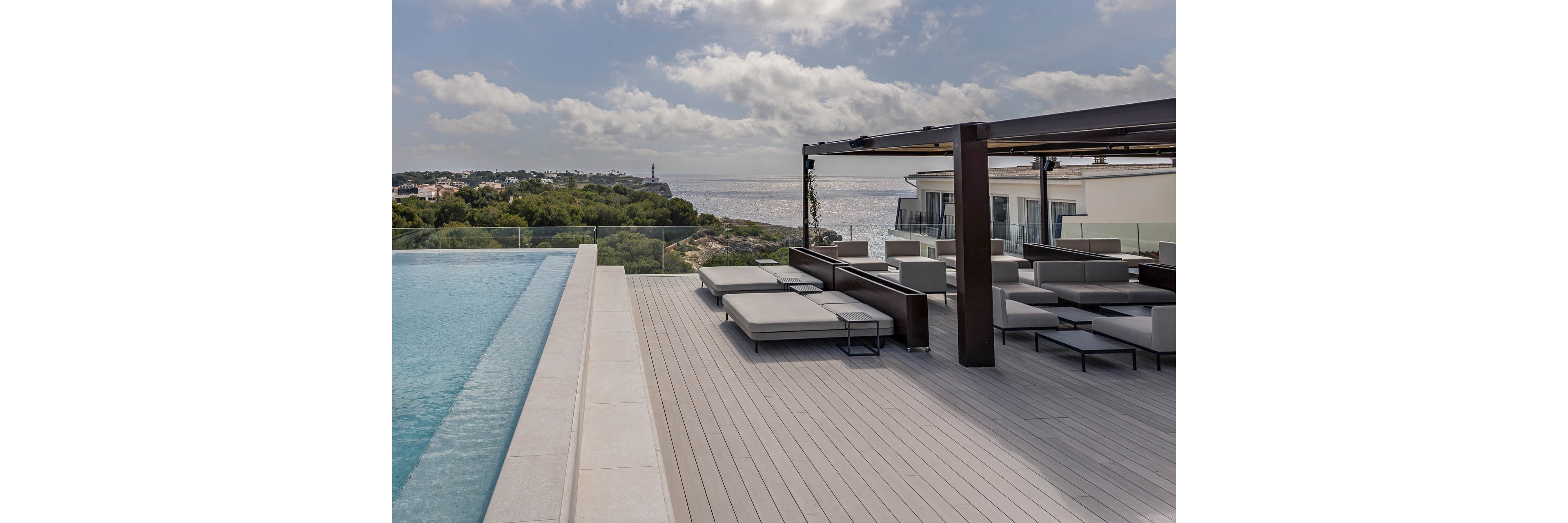 A contemporary rooftop terrace features an expansive infinity pool overlooking a coastal landscape. Sleek gray loungers and modular sofas sit beneath a dark wooden pergola, creating a stylish outdoor lounge area. Glass railings frame the deck, offering unobstructed views of the sea and distant shoreline buildings. Soft clouds and calm water contribute to a relaxed, luxurious resort atmosphere.