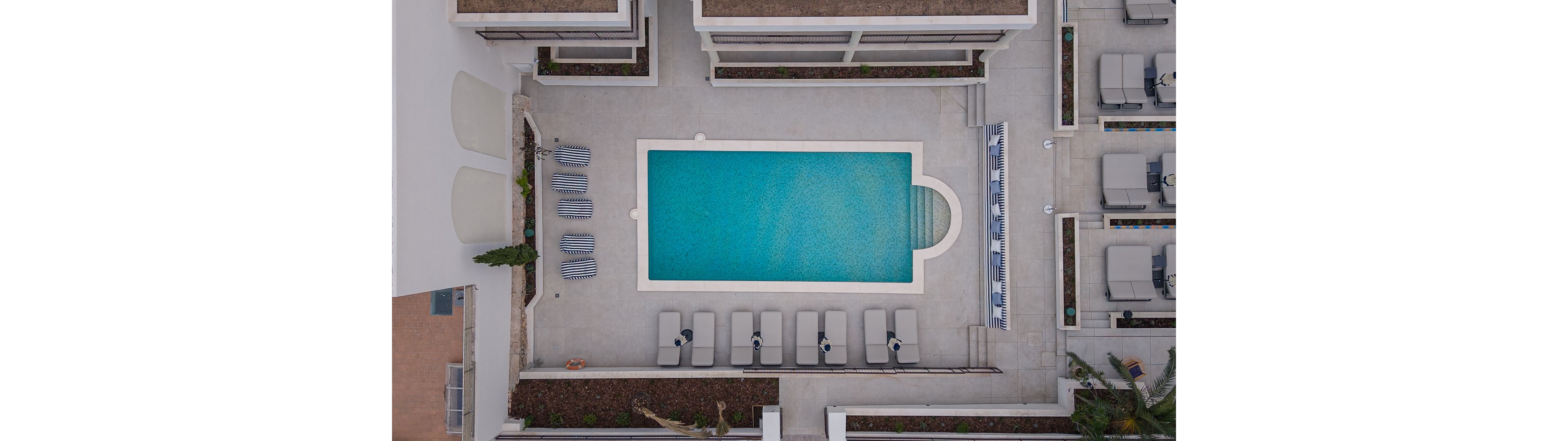 A rectangular outdoor swimming pool is photographed from directly above in a modern hotel or resort courtyard. Lounge chairs and striped sunbeds are neatly arranged around the pool on a light-colored deck. The water appears clear blue, with steps visible at one end of the pool. Surrounding planters and building structures frame the scene, emphasizing symmetry and order.