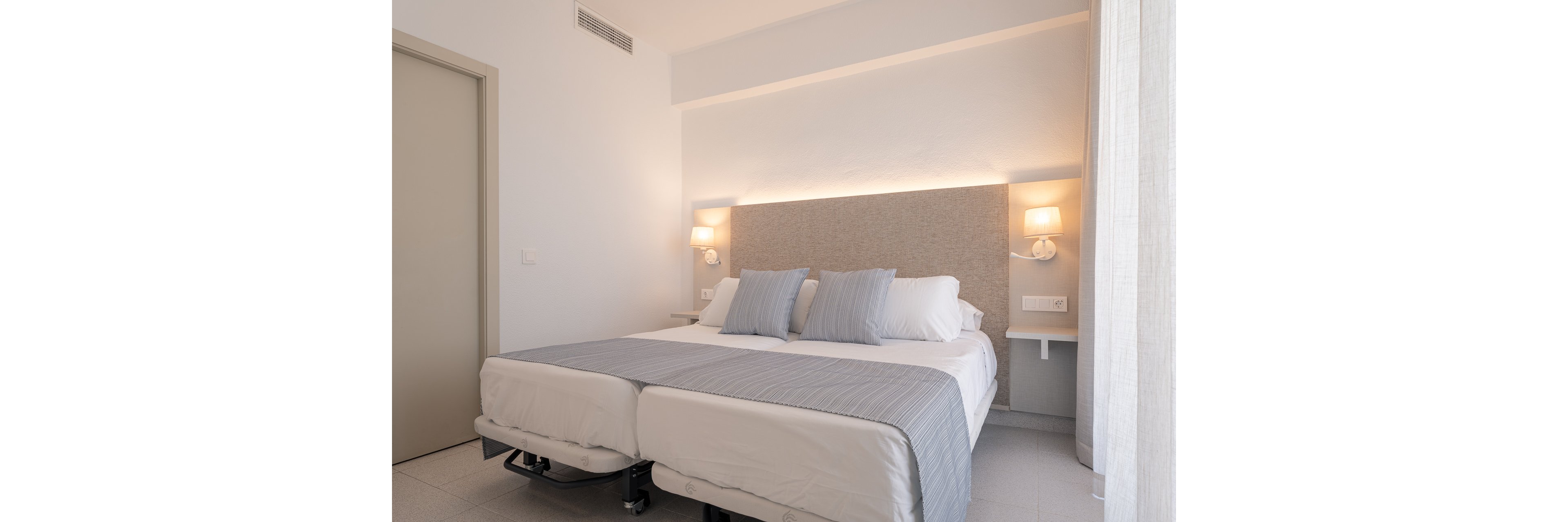 A bright modern bedroom features two twin beds pushed together, dressed in white linens and light gray throws. Three matching gray pillows rest against a padded headboard that spans the wall. Warm wall-mounted lamps on either side of the bed provide soft ambient lighting. Minimalist white furniture and neutral tones create a clean, contemporary hotel-like atmosphere.