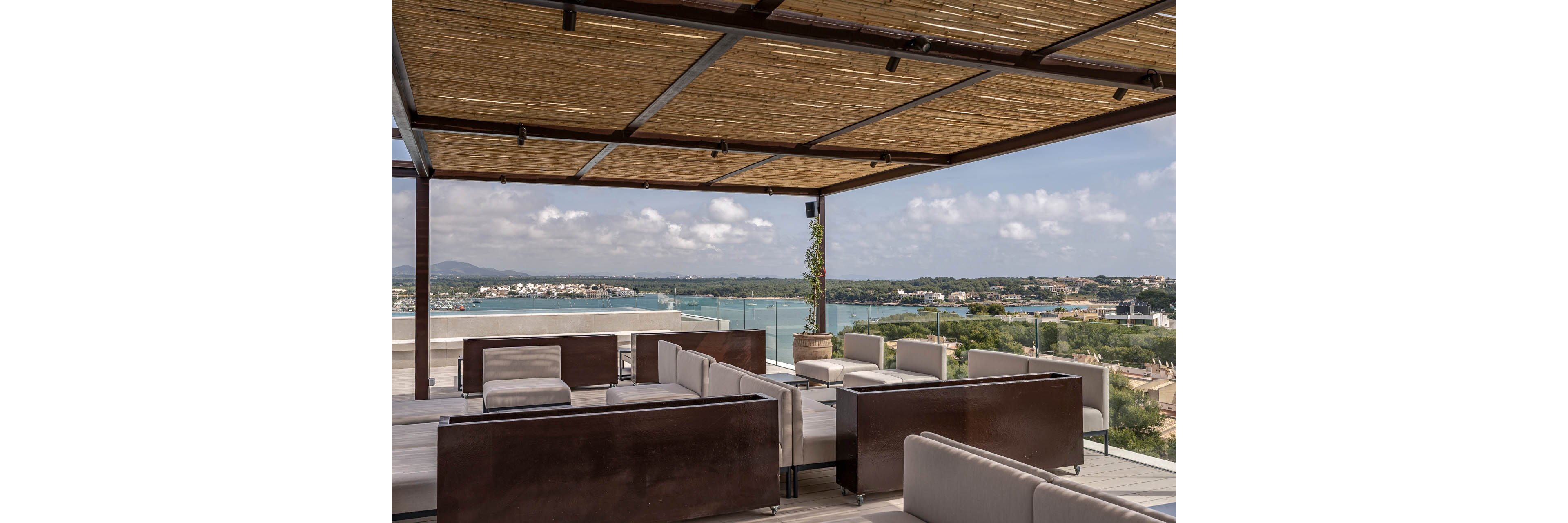 A contemporary rooftop lounge features sleek modular sofas and low tables arranged beneath a wooden pergola. The open terrace offers expansive views of a coastal bay, with calm blue water and a distant shoreline dotted with buildings. Glass railings frame the edge of the terrace, maintaining unobstructed vistas of the landscape. Neutral tones and minimalist design create a relaxed, upscale outdoor atmosphere.