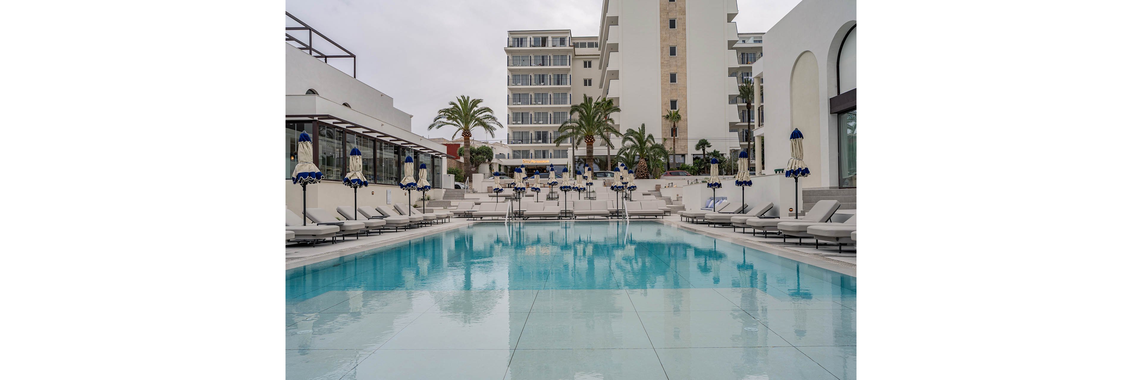 A large, clear swimming pool sits at the center of a modern resort complex. White lounge chairs and closed umbrellas line both sides of the pool, creating a symmetrical and orderly scene. Multi-story hotel buildings and palm trees rise in the background under an overcast sky. The overall atmosphere feels calm, minimal, and luxurious, emphasizing relaxation and leisure.