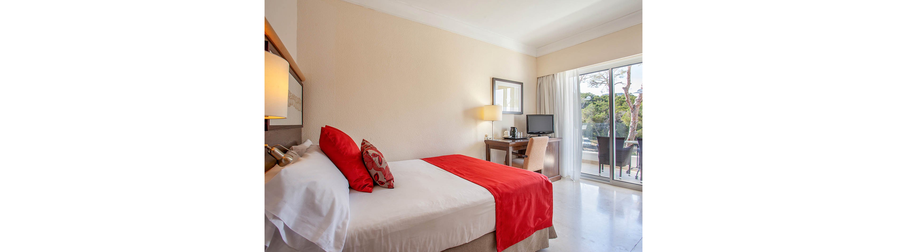 A neatly made double bed with white linens and red accents is the focal point of this hotel room. The space includes a wooden desk with a chair, table lamp, and a flat-screen television. Large sliding glass doors open to a balcony with outdoor seating and a view of trees, allowing natural light to fill the room. The overall atmosphere is clean, modern, and inviting, suitable for vacation or business travel.