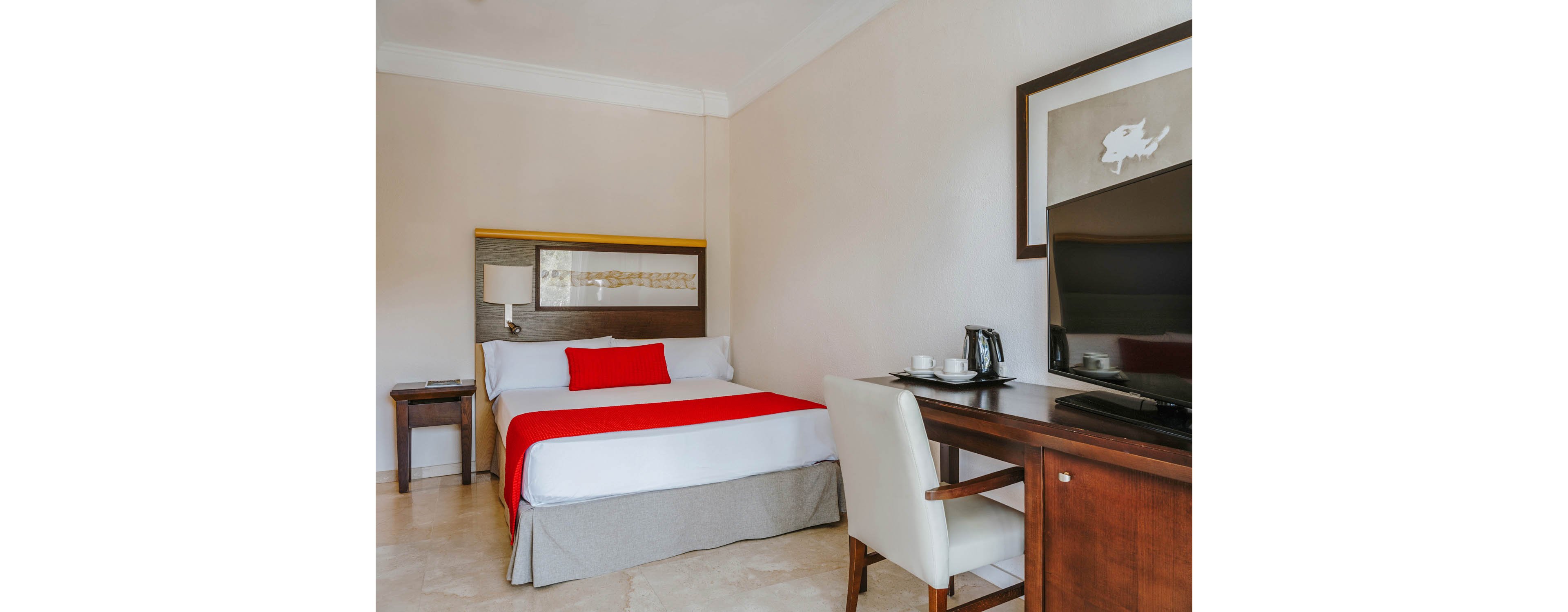 A neatly made bed with white linens and a bright red runner and pillow is the focal point of this modern hotel room. The space features neutral walls, a wooden headboard, and a framed artwork above the bed. To the right, a wooden desk holds a flat-screen television, cups, and a coffee maker, with a white upholstered chair tucked underneath. A small bedside table with a lamp completes the simple, contemporary decor.