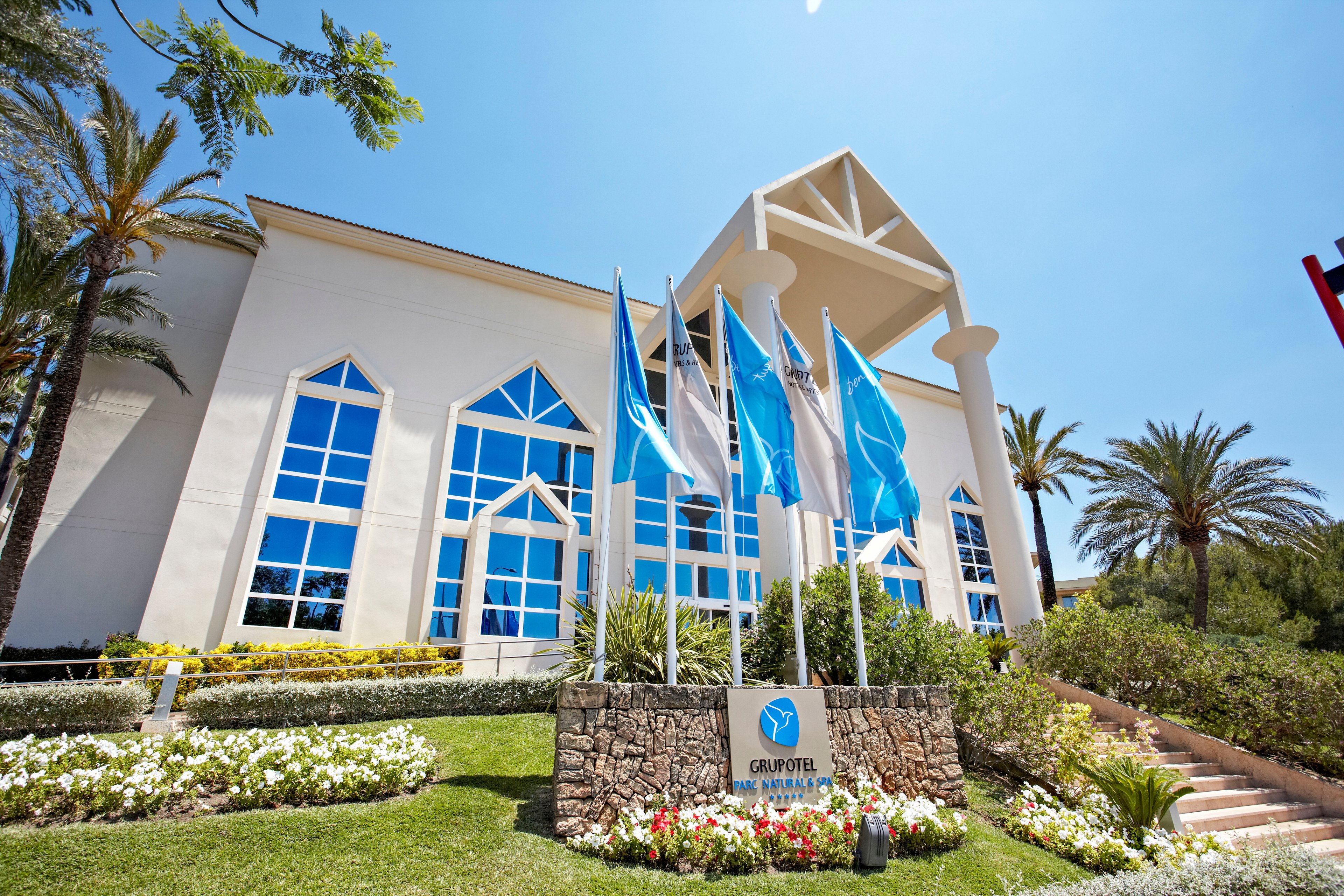 A contemporary white hotel building stands behind a row of tall flagpoles with blue and white flags. The structure features large geometric windows and a prominent triangular roof detail. Lush green lawns, colorful flowerbeds, and palm trees surround the entrance, creating a tropical atmosphere. Bright sunlight and a clear blue sky emphasize the clean, inviting resort setting.