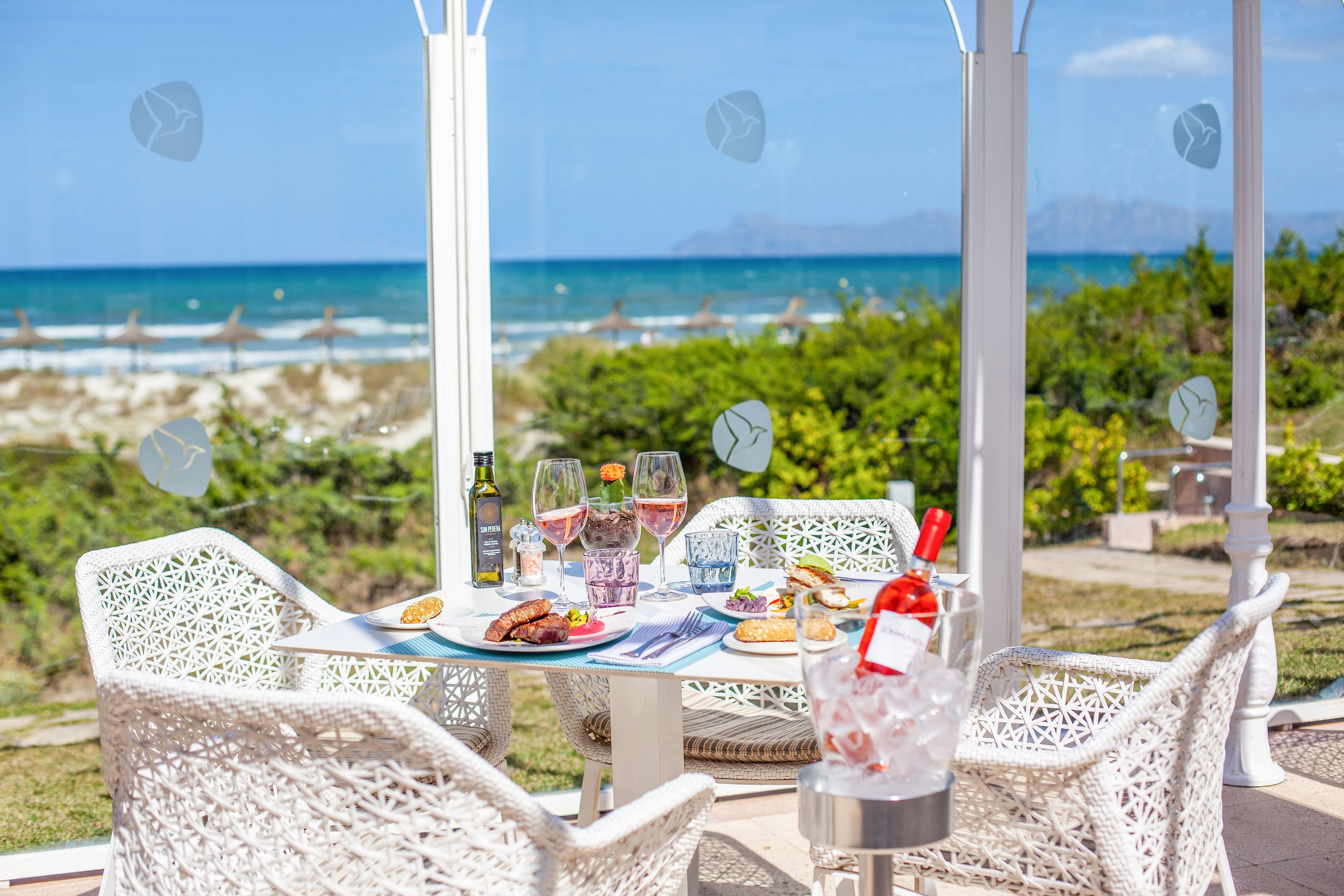 A stylish outdoor dining table is set with plates of food and several glasses of rosé wine. White woven chairs surround the table under a light pergola structure overlooking a sandy beach and blue ocean. A bottle of wine chills in an ice bucket beside the table, while another bottle and water glasses sit near the plates. The scene is bright and sunny, with clear skies and a relaxed coastal atmosphere.