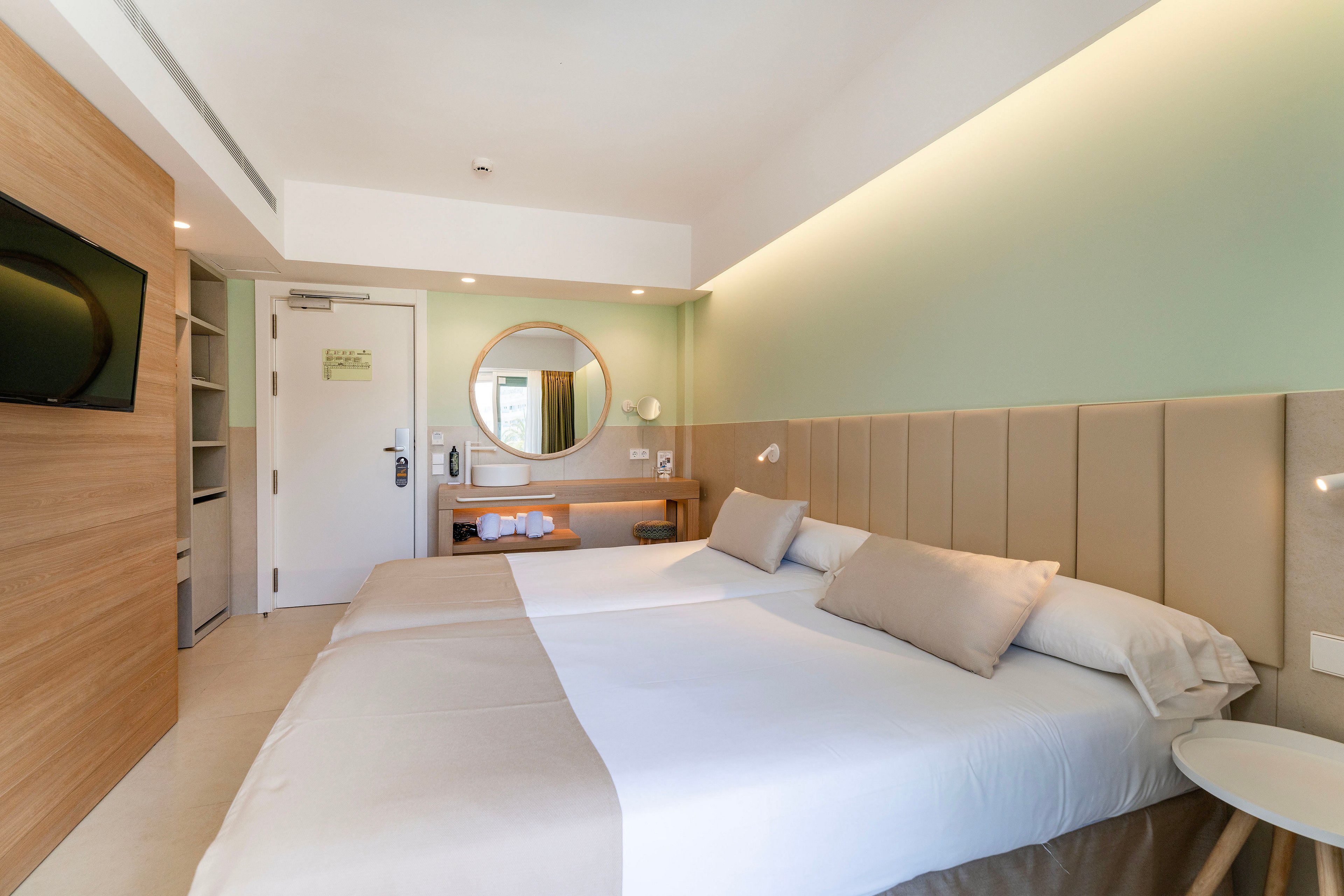 A contemporary hotel room features two neatly made twin beds with neutral bedding and accent pillows. The space is decorated in soft beige and pastel green tones, with a padded headboard running along the wall. A round mirror hangs above a wooden desk, and a flat-screen TV is mounted opposite the beds. Natural light brightens the minimalist interior, creating a clean and inviting atmosphere.