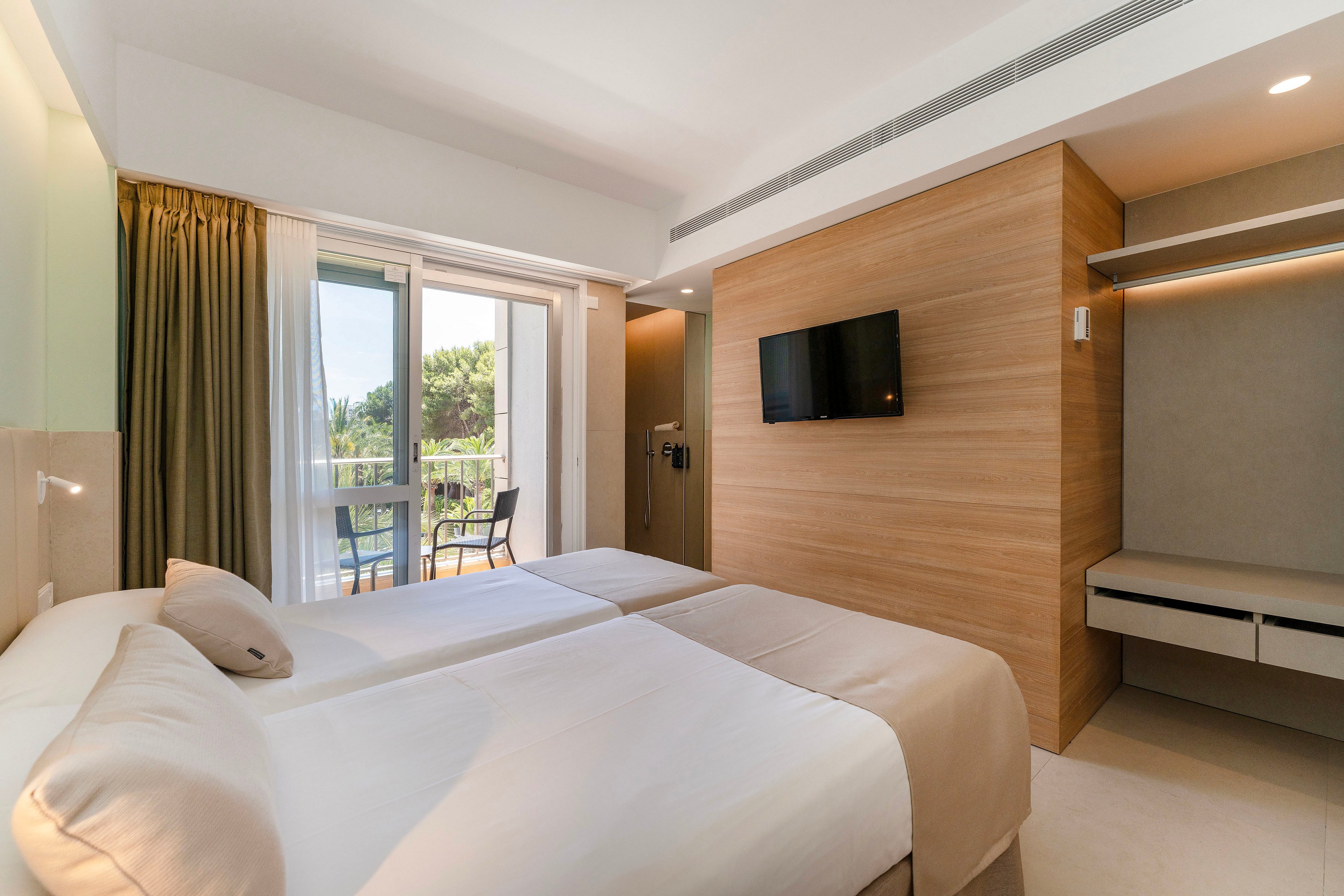 A contemporary hotel room features two neatly made twin beds with neutral bedding. A wall-mounted flat-screen television is set against a light wood accent wall, opposite a minimalist desk area. Large glass doors open onto a balcony with outdoor chairs, overlooking green trees. Soft natural light and warm tones create a calm, inviting atmosphere suitable for relaxation or business travel.