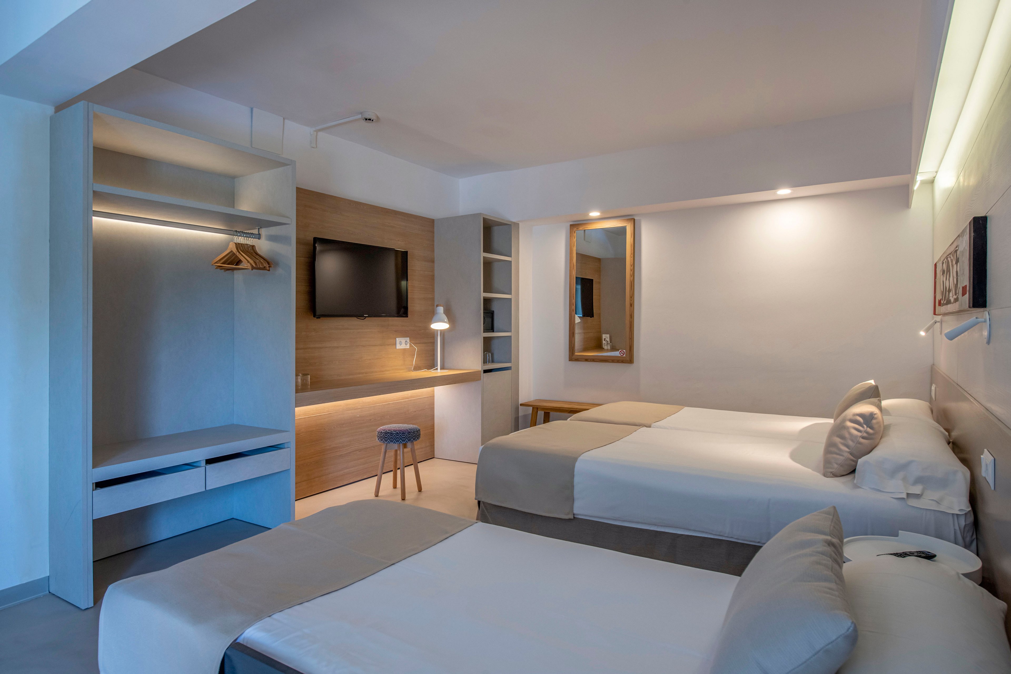 Contemporary hotel room interior featuring two neatly made beds with neutral bedding and accent pillows. A wall-mounted flat-screen television faces the beds above a long wooden desk with a stool and task lamp. Open closet space with hangers and built-in shelving is visible to the side, along with a large mirror and soft ambient lighting. The overall design is minimalist, with clean lines and a calm, muted color palette.