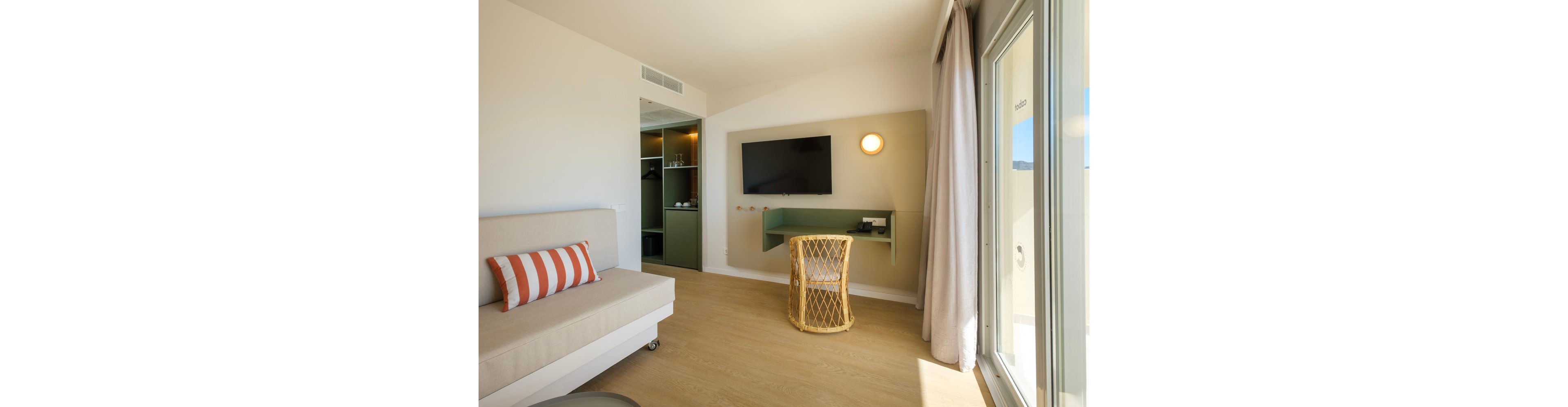A bright modern hotel room features a wall-mounted flat screen television opposite a light-colored sofa with a striped cushion. A small woven chair sits at a minimalist green desk beneath the TV, with a round wall light providing warm illumination. Built-in shelving and a wardrobe are visible in the open closet area, adding storage space. Large windows with light curtains allow natural daylight to fill the room, creating an airy and inviting atmosphere.