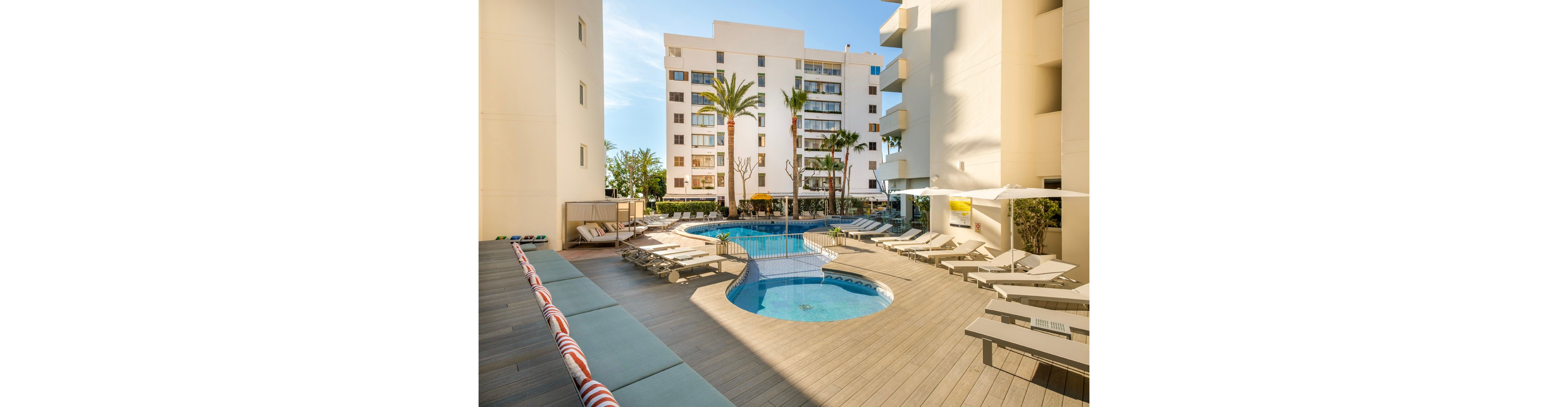 A contemporary hotel courtyard features a central outdoor swimming pool surrounded by sun loungers and umbrellas. The pool has a curved shape with a small raised circular section and steps leading into the water. Tall residential-style buildings and palm trees frame the space, creating an urban resort atmosphere. Cushioned bench seating with striped pillows lines one side of the deck, emphasizing comfort and relaxation.