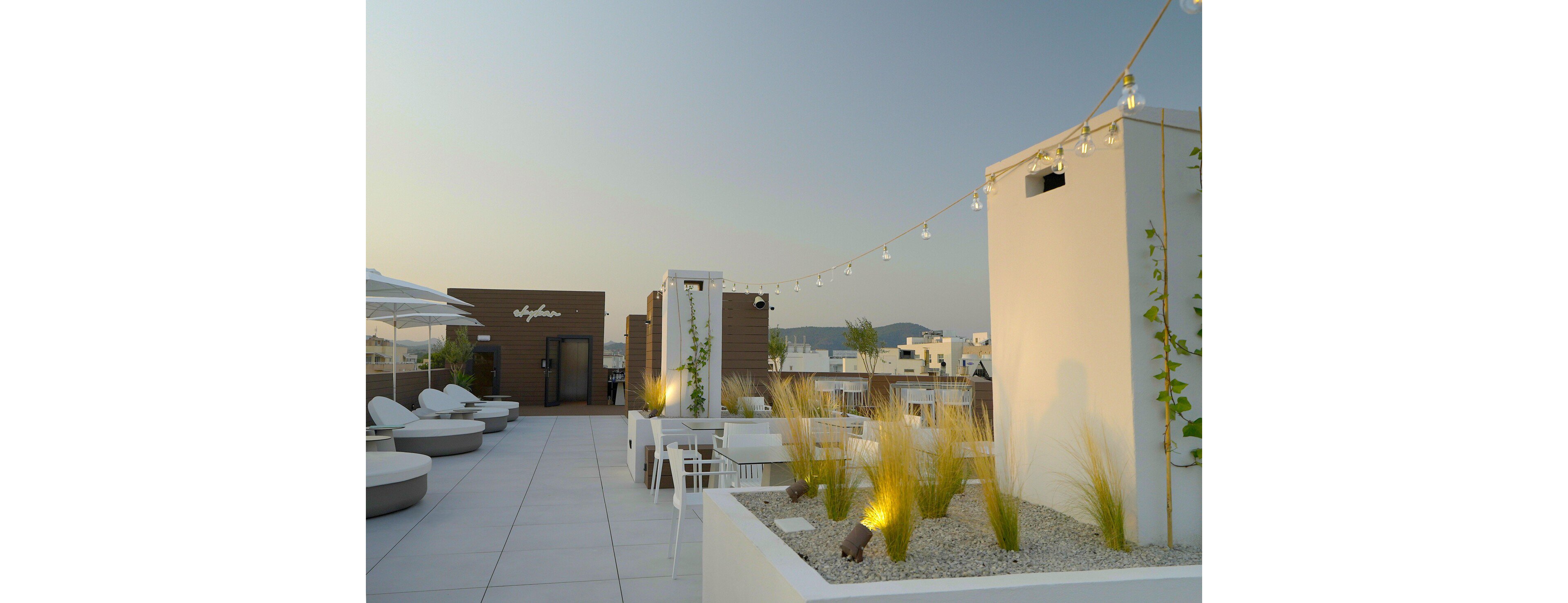 A contemporary rooftop terrace features minimalist white seating and planters filled with ornamental grasses. Warm string lights are suspended above the space, creating a cozy atmosphere at dusk. Clean architectural lines and neutral tones emphasize the modern design. Surrounding buildings and distant hills are visible under a clear evening sky.