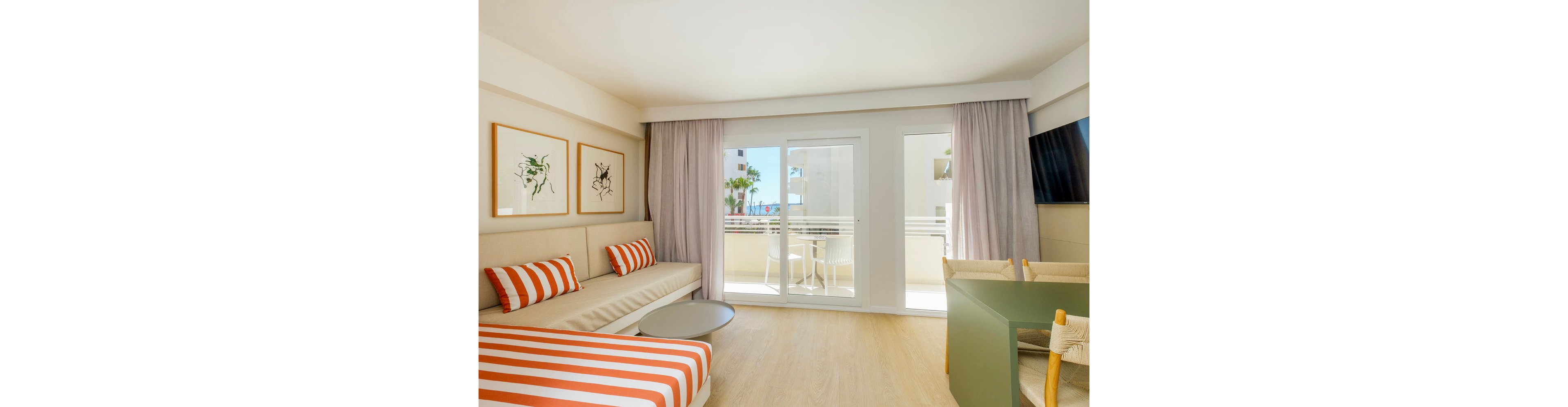 A modern hotel room features a striped daybed and matching cushions in warm orange and white tones. Large sliding glass doors open onto a sunny balcony with outdoor chairs and a table. Light wood flooring, neutral walls, and soft curtains create an airy, minimalist atmosphere. Framed artwork and a wall-mounted television complete the contemporary interior design.