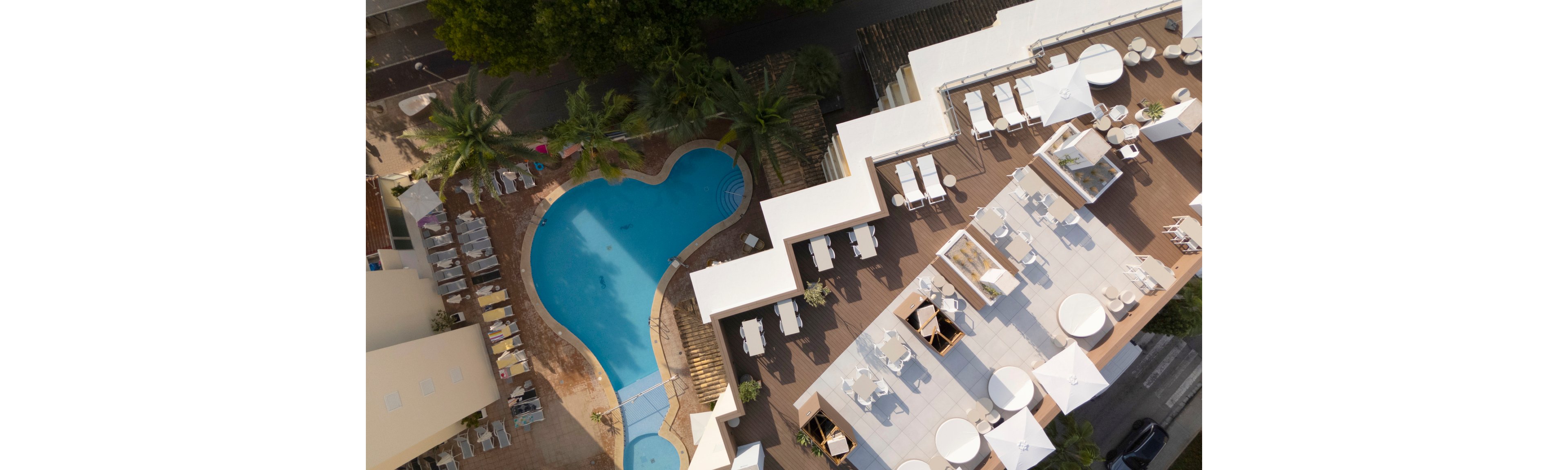 An aerial view shows a modern hotel complex with a large, irregularly shaped swimming pool. Sun loungers and umbrellas are arranged neatly around the pool and on an adjacent rooftop terrace. The scene includes palm trees, shaded seating areas, and clean geometric lines of the building structures. Warm lighting and soft shadows suggest a relaxed, resort-style atmosphere in a tropical or coastal setting.