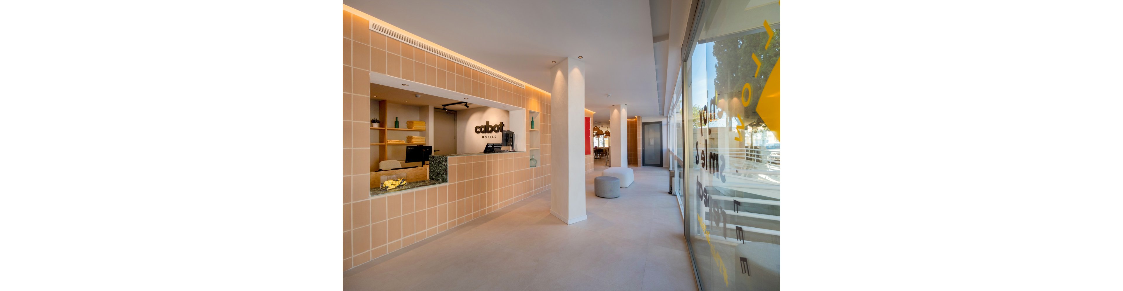 A contemporary hotel reception area features a tiled front desk with a computer and decorative shelving behind it. The space is designed with warm beige tones, clean lines, and recessed lighting that creates a welcoming atmosphere. Large glass windows on the right allow natural light to fill the lobby and offer a view outside. In the background, soft seating and a corridor lead further into the hotel interior.