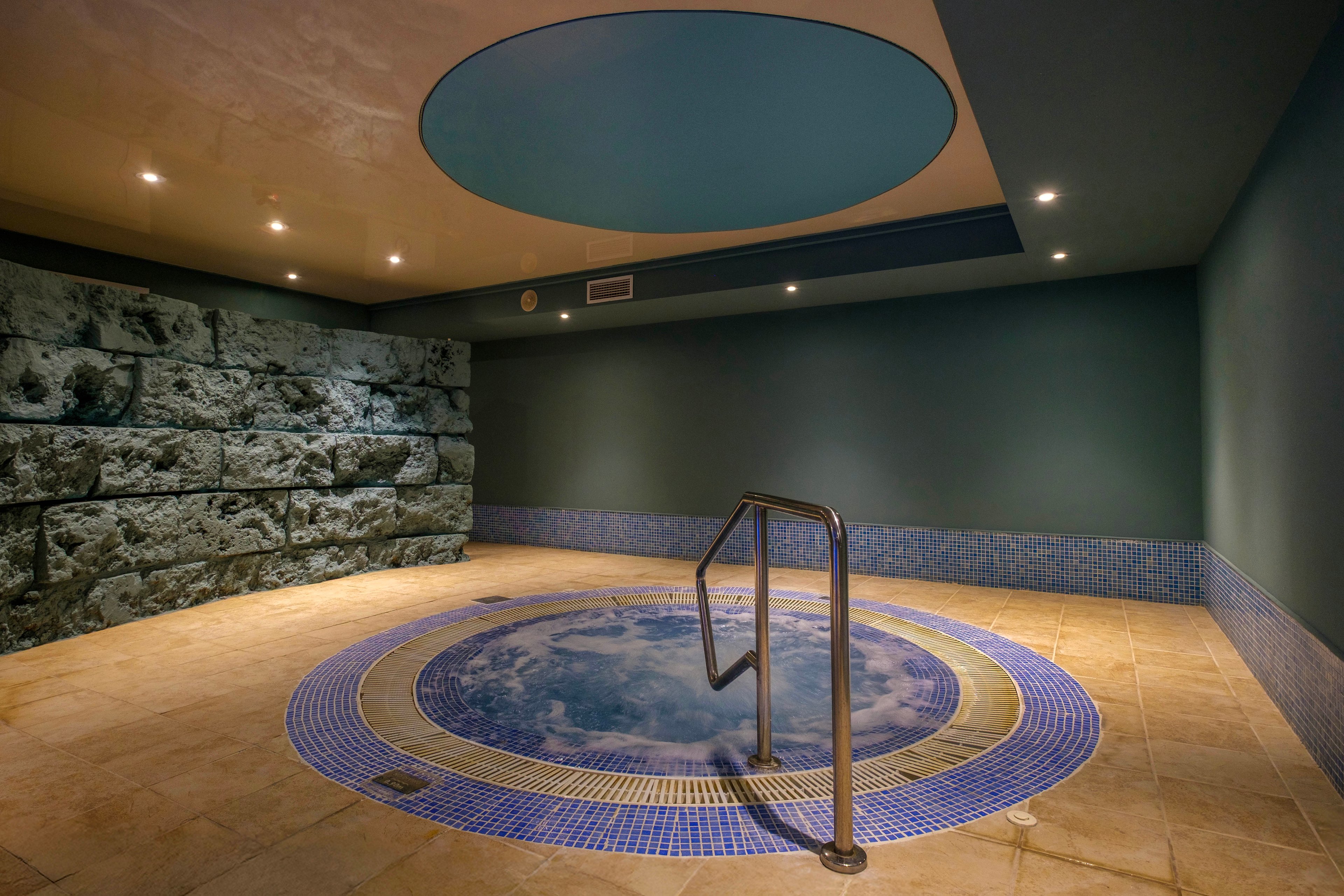 An indoor spa room features a small circular pool set into a tiled floor. The pool is bordered by blue mosaic tiles and includes a stainless steel handrail for safe entry. One wall is finished with textured stone blocks, while the others are smooth and painted in muted tones. Recessed ceiling lights create a calm, softly lit atmosphere suitable for relaxation.