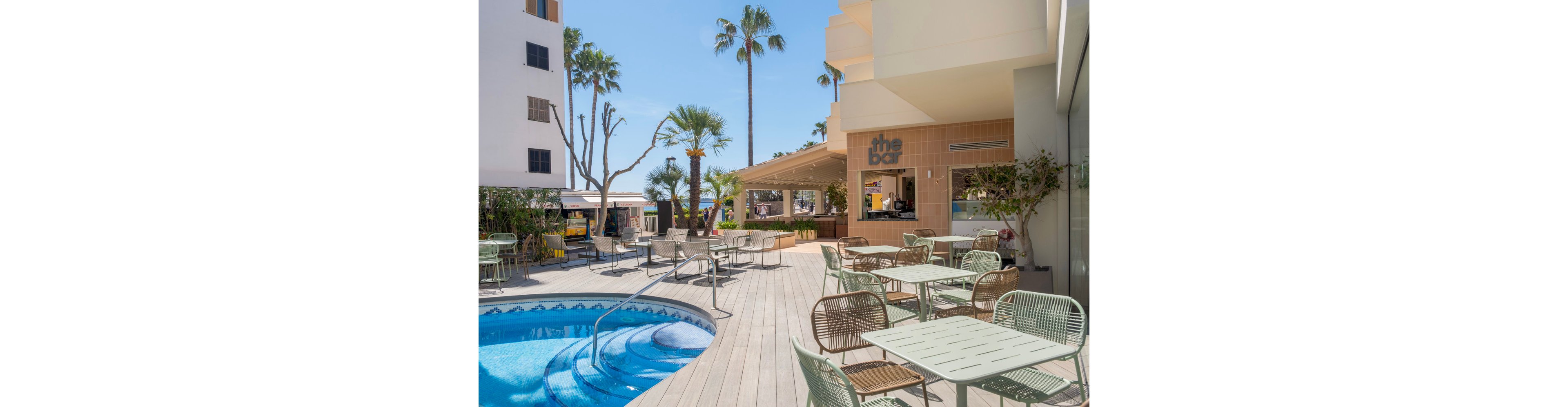 An outdoor bar terrace overlooks a curved swimming pool surrounded by a wooden deck. Metal and wicker chairs with matching tables are arranged neatly around the pool area. Palm trees and modern buildings frame the sunny, resort-like setting. A sign reading "the bar" is visible above the open counter, suggesting a relaxed place for drinks and socializing.