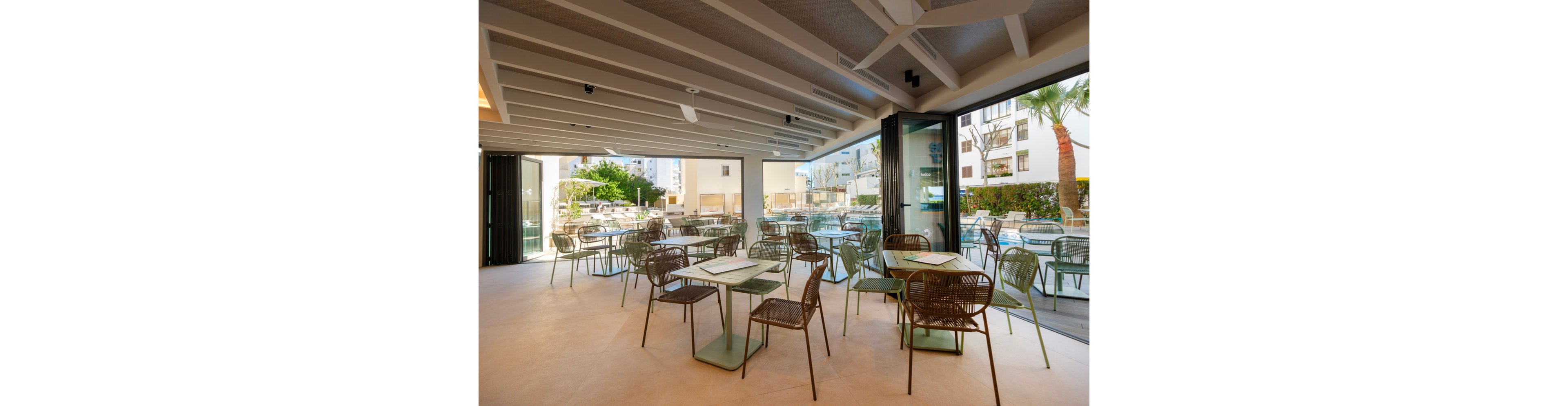 A contemporary café or restaurant features neatly arranged tables and metal chairs in a bright, open-plan interior. Large glass doors open onto an outdoor terrace with additional seating beside a swimming pool. Sunlight streams in, highlighting the clean lines, neutral flooring, and minimalist décor. The surrounding buildings and palm trees create a relaxed, resort-like atmosphere.