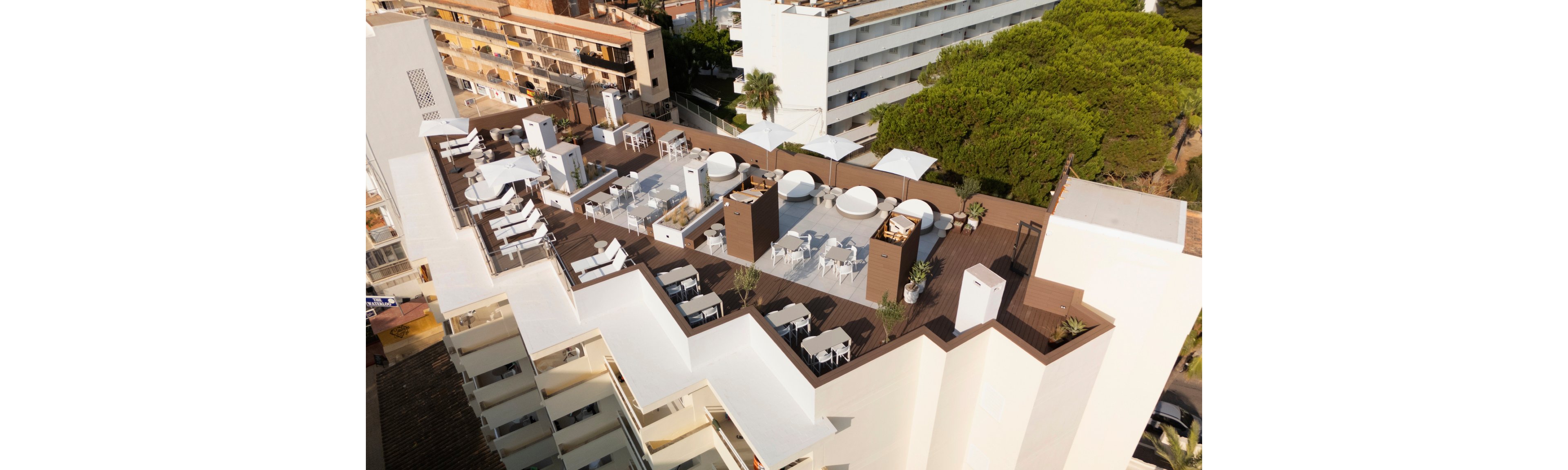 An aerial view shows a modern rooftop terrace on top of a multi-story building. The deck features lounge chairs, round daybeds, tables, and umbrellas arranged in several seating zones. White and brown tones dominate the design, contrasting with the surrounding urban buildings and nearby trees. The scene suggests a relaxed outdoor social or leisure area in a city setting.