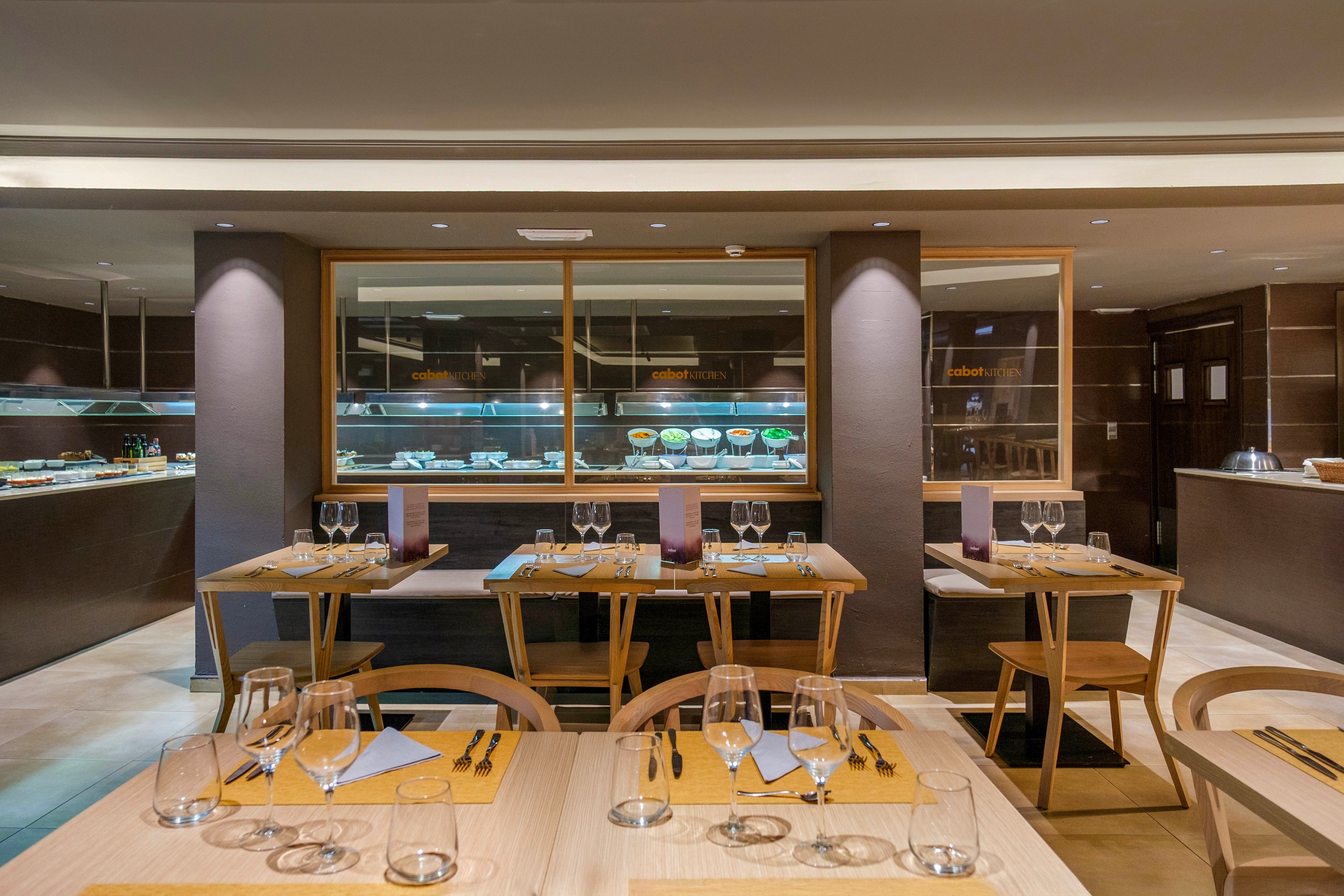 A contemporary restaurant dining room is set with neatly arranged wooden tables and chairs. Each table is prepared with wine glasses, folded napkins, and cutlery, ready for guests. In the background, a glass-fronted buffet showcases various dishes under warm lighting. The overall design features clean lines, neutral tones, and a sophisticated, minimalist aesthetic.