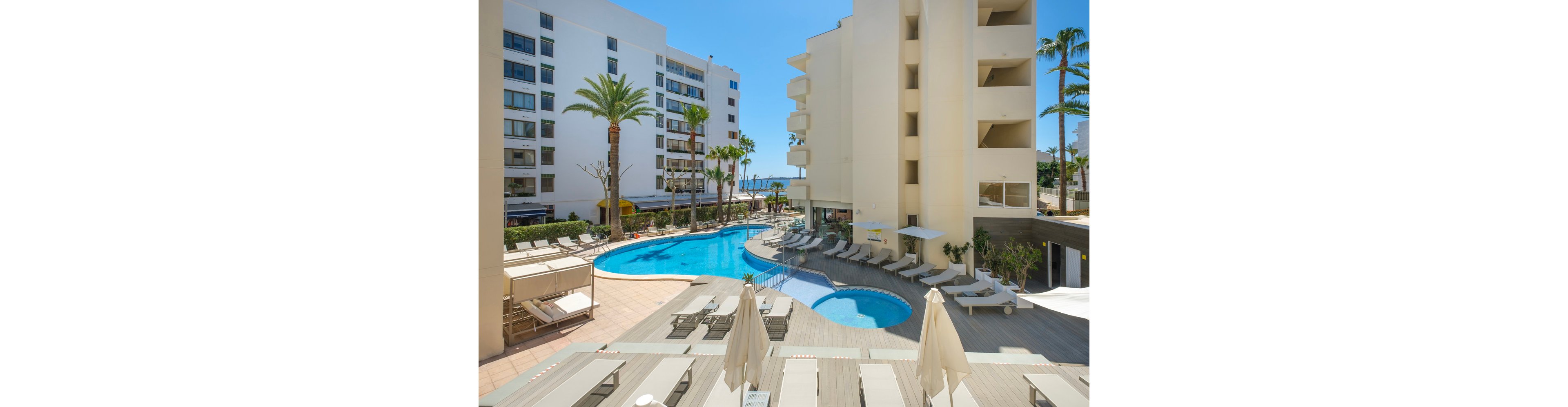 A bright hotel courtyard features a curving outdoor swimming pool surrounded by white sun loungers and umbrellas. Modern multi-story buildings frame the scene, with balconies overlooking the pool area. Palm trees and clear blue skies suggest a warm coastal or resort destination. The overall atmosphere is relaxed and inviting, ideal for sunbathing and leisure by the water.