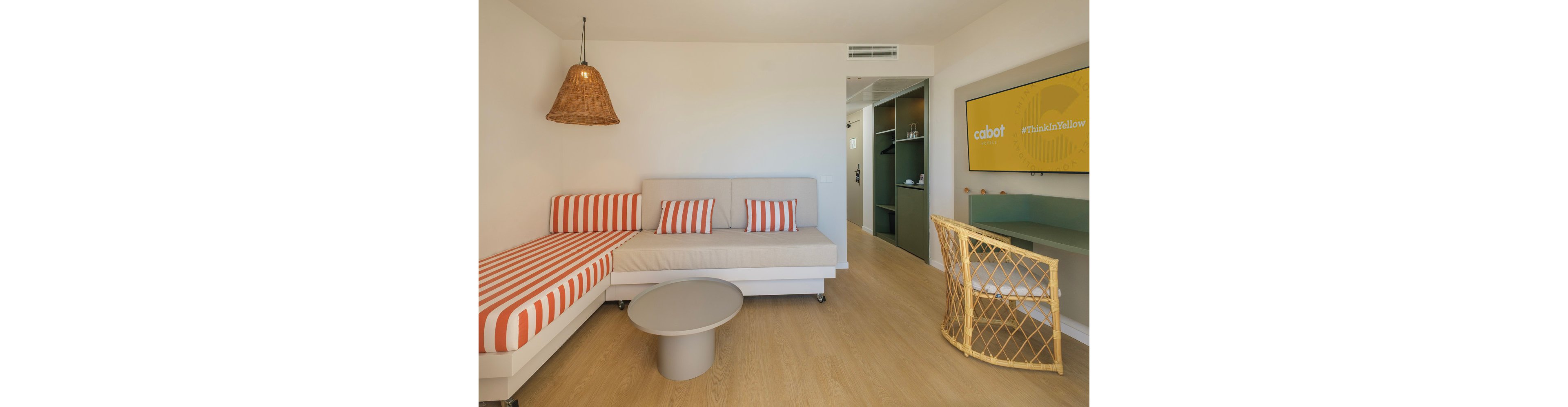 A contemporary hotel room features a white sofa and a daybed with orange and white striped cushions. A round gray coffee table sits on a light wood floor, adding to the minimalist decor. A wicker pendant light and matching wicker chair bring warmth and texture to the space. On the wall, a yellow screen displays the word cabot and the hashtag ThinkInYellow, reinforcing the room’s bright, cheerful theme.