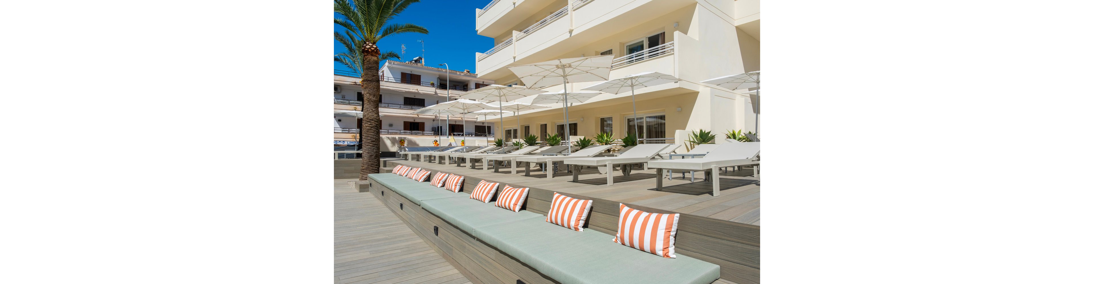 A contemporary hotel terrace features a long cushioned bench with striped orange and white pillows in the foreground. Behind it, rows of white sun loungers and large umbrellas are arranged neatly on a wooden deck. The building’s balconies overlook the terrace, and a palm tree adds a touch of greenery. Bright daylight and clear skies create a relaxed, vacation atmosphere.