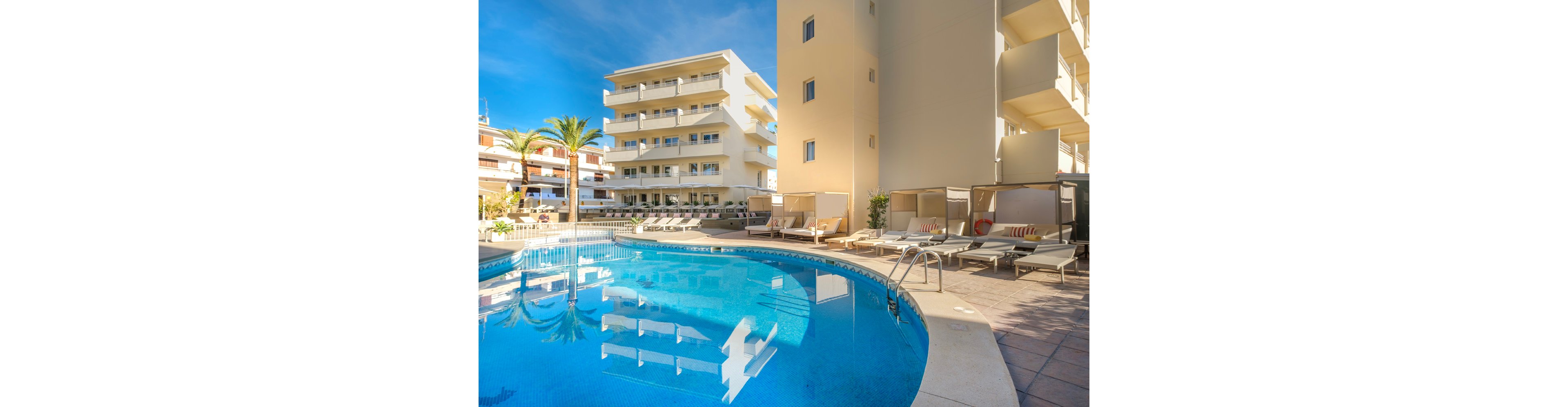 A bright hotel courtyard features a curved blue swimming pool surrounded by modern apartment-style buildings. Sun loungers and shaded cabanas line the tiled pool deck, suggesting a relaxing resort atmosphere. A tall palm tree and clear blue sky enhance the warm, coastal feel of the scene. The overall setting appears calm and inviting, ideal for vacation or leisure time.