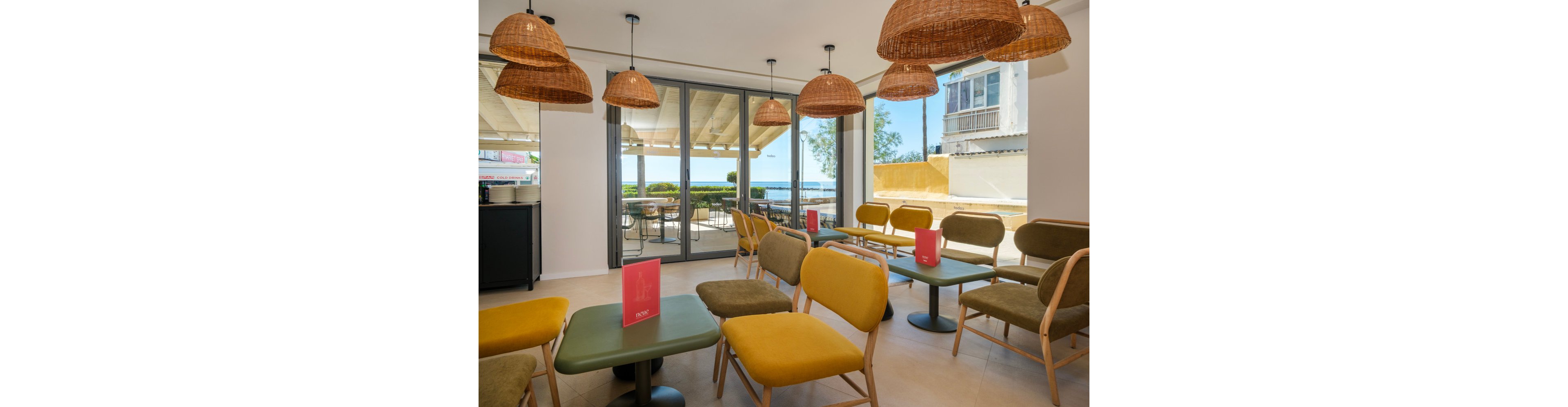 A bright modern café interior features mustard-yellow chairs and green tables arranged in neat rows. Large glass doors open to an outdoor terrace with additional seating and a view of the sea. Woven wicker pendant lamps hang from the ceiling, adding warmth and texture to the minimalist space. Pink table menus stand on several tables, providing a subtle pop of color against the neutral walls and flooring.