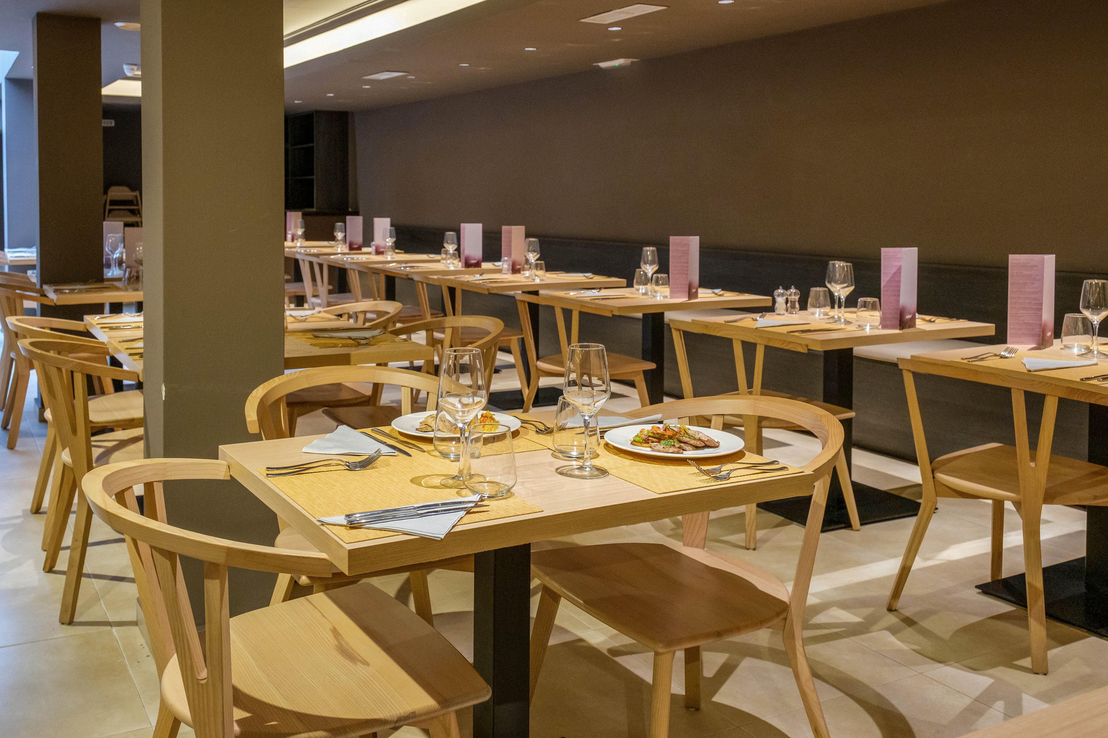 A contemporary restaurant interior features light wooden tables and matching chairs neatly arranged in rows. Each table is set with wine glasses, folded napkins, cutlery, and menus, creating a ready-for-service atmosphere. Warm, indirect lighting and neutral wall colors contribute to a calm, minimalist aesthetic. A single plated dish is visible in the foreground, adding a subtle touch of color and focus.