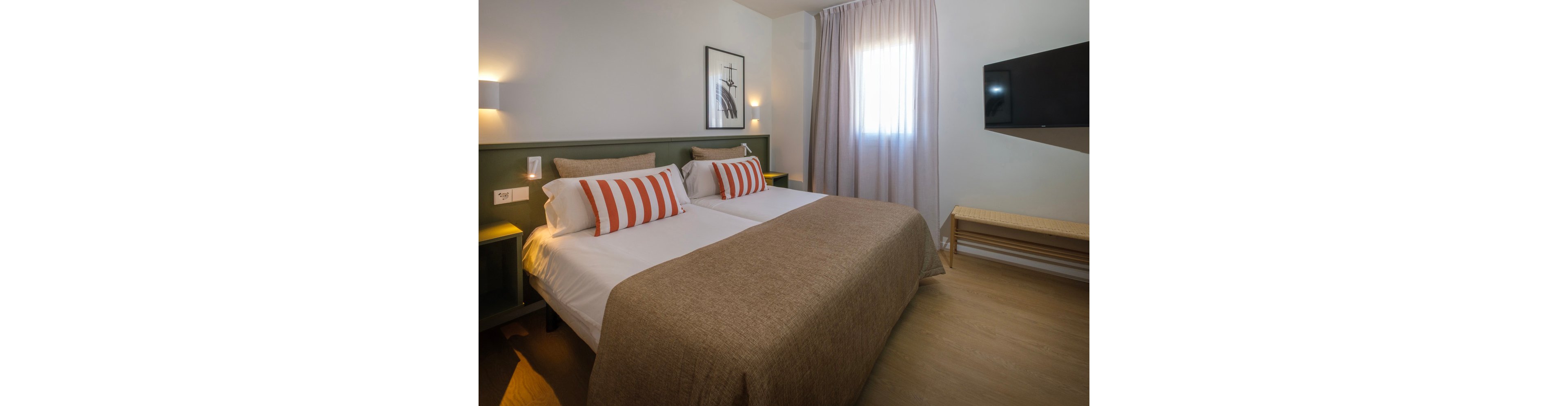 A contemporary hotel room features two twin beds pushed together, topped with brown bedspreads and striped accent pillows. Soft wall lighting and a framed artwork create a cozy, minimalist atmosphere. A window with sheer curtains lets in natural light, while a flat-screen television is mounted on the wall opposite the beds. A simple wooden bench and neutral color palette contribute to the clean, modern design.