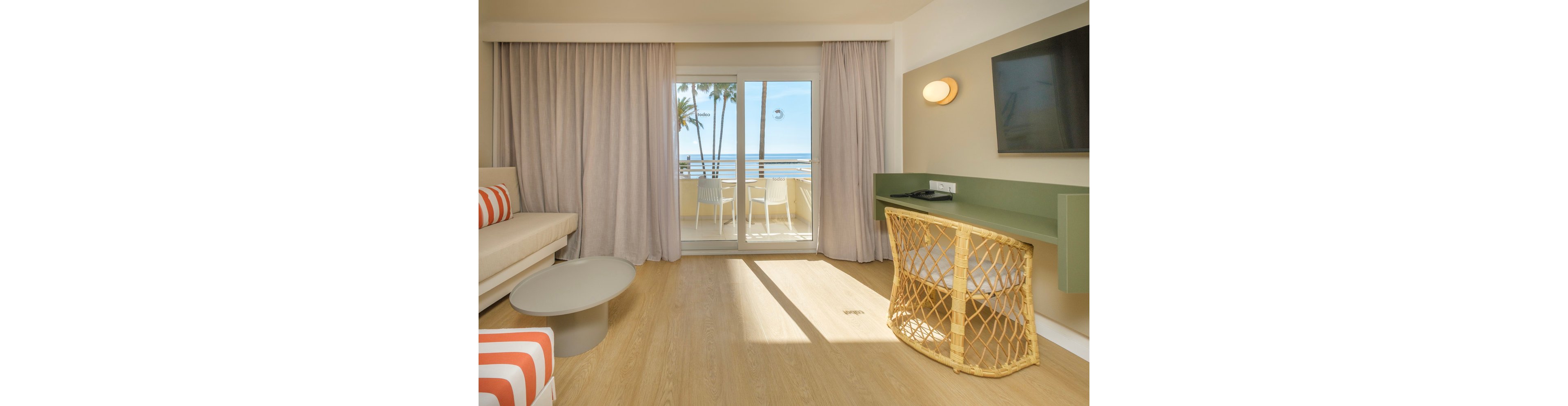 A bright modern hotel room features a sofa, low round table, and wall-mounted television. Light wood flooring and neutral walls create a clean, minimalist interior. Sliding glass doors open to a balcony with white chairs and a view of palm trees and the ocean. Soft curtains and warm lighting contribute to a relaxed, coastal atmosphere.