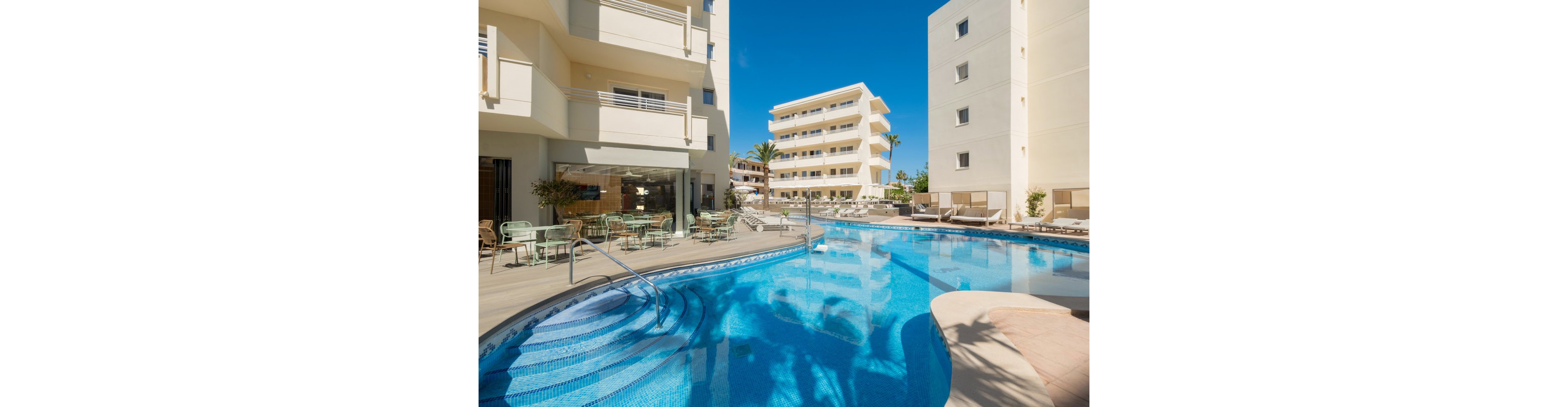 A bright outdoor swimming pool sits in the courtyard of a modern hotel complex. Multi-story white buildings surround the pool, featuring balconies and large windows. Sunlight reflects off the clear blue water, and metal handrails lead into the shallow steps. Patio tables and chairs line the poolside area, creating a relaxed resort atmosphere under a clear blue sky.