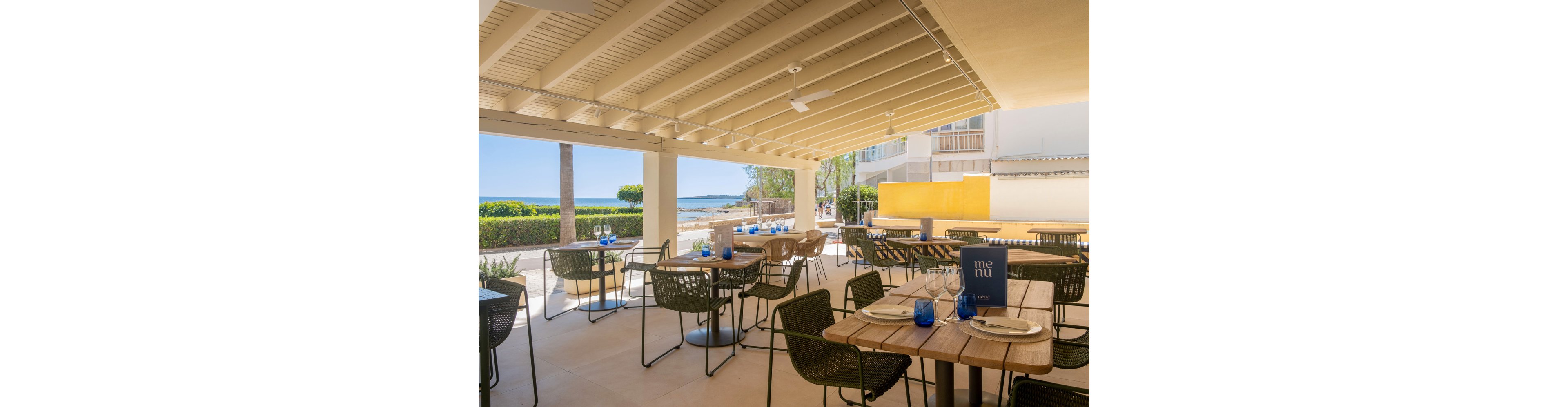 An open-air seaside restaurant terrace features neatly arranged wooden tables and woven chairs. Place settings with plates, cutlery, and blue glasses are prepared for guests under a slatted pergola roof. A blue menu stands on one of the tables, while the bright coastline and calm sea are visible in the background. The scene suggests a relaxed, sunny dining experience by the water.