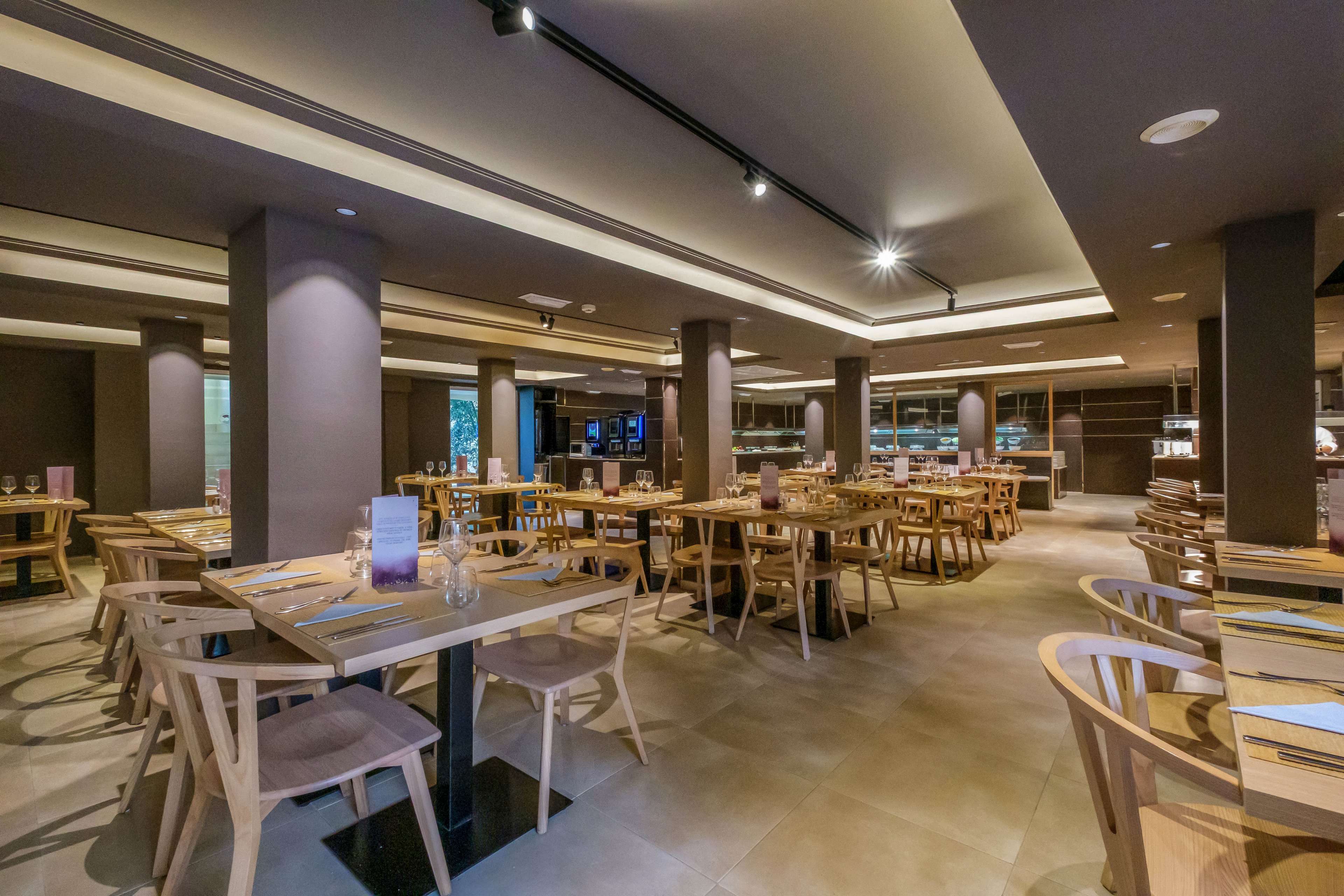 Spacious contemporary restaurant interior with neatly arranged wooden tables and chairs. The dining room is empty, with place settings and menus prepared for guests. Warm ceiling lights and neutral colors create a cozy, inviting atmosphere. Large windows and open layout suggest a casual yet stylish dining experience.