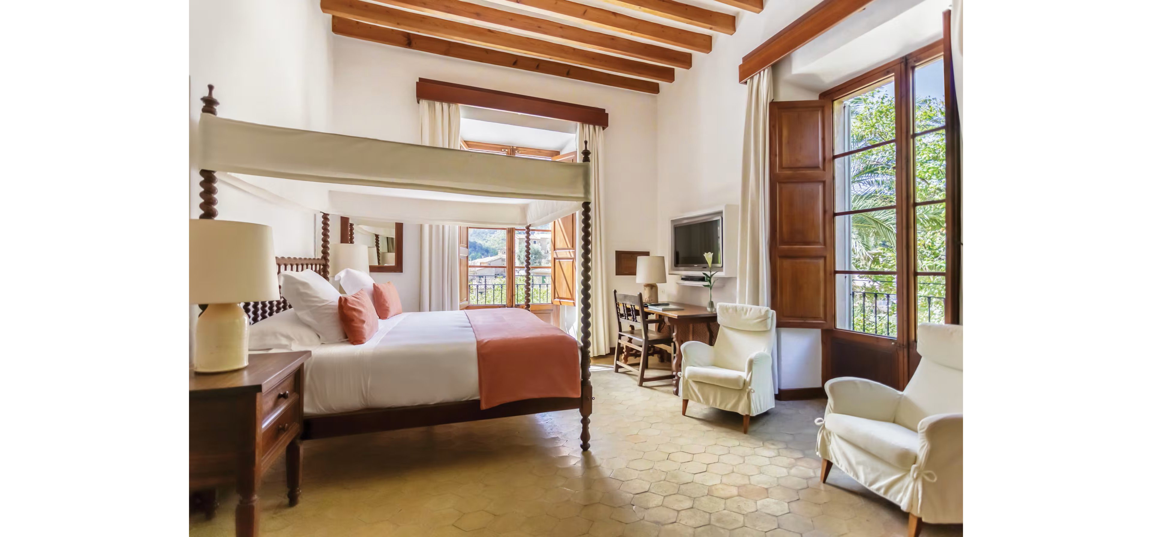 A spacious, sunlit bedroom features a four-poster bed with white linens and soft terracotta accents. The room includes wooden furniture, a writing desk with a lamp, and two upholstered armchairs. Large wooden-framed windows and doors open to a balcony with views of greenery and distant hills. Exposed wooden ceiling beams and patterned floor tiles enhance the warm, Mediterranean-inspired atmosphere.