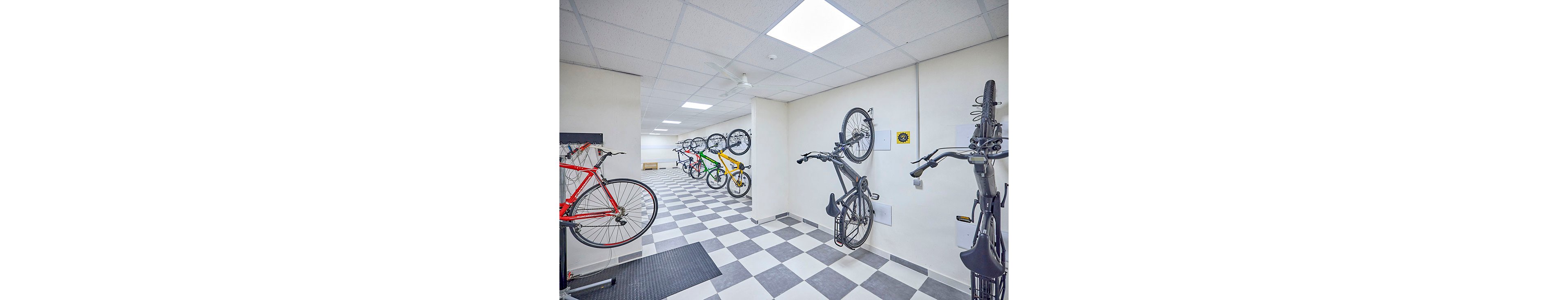 A clean, modern indoor room features multiple bicycles stored on wall-mounted racks. The space has white walls, a checkered gray and white tile floor, and bright ceiling lights. Several bikes are hung vertically along the right wall, while others are mounted horizontally on the left and back walls. The organized layout suggests a secure storage or maintenance area for cyclists.