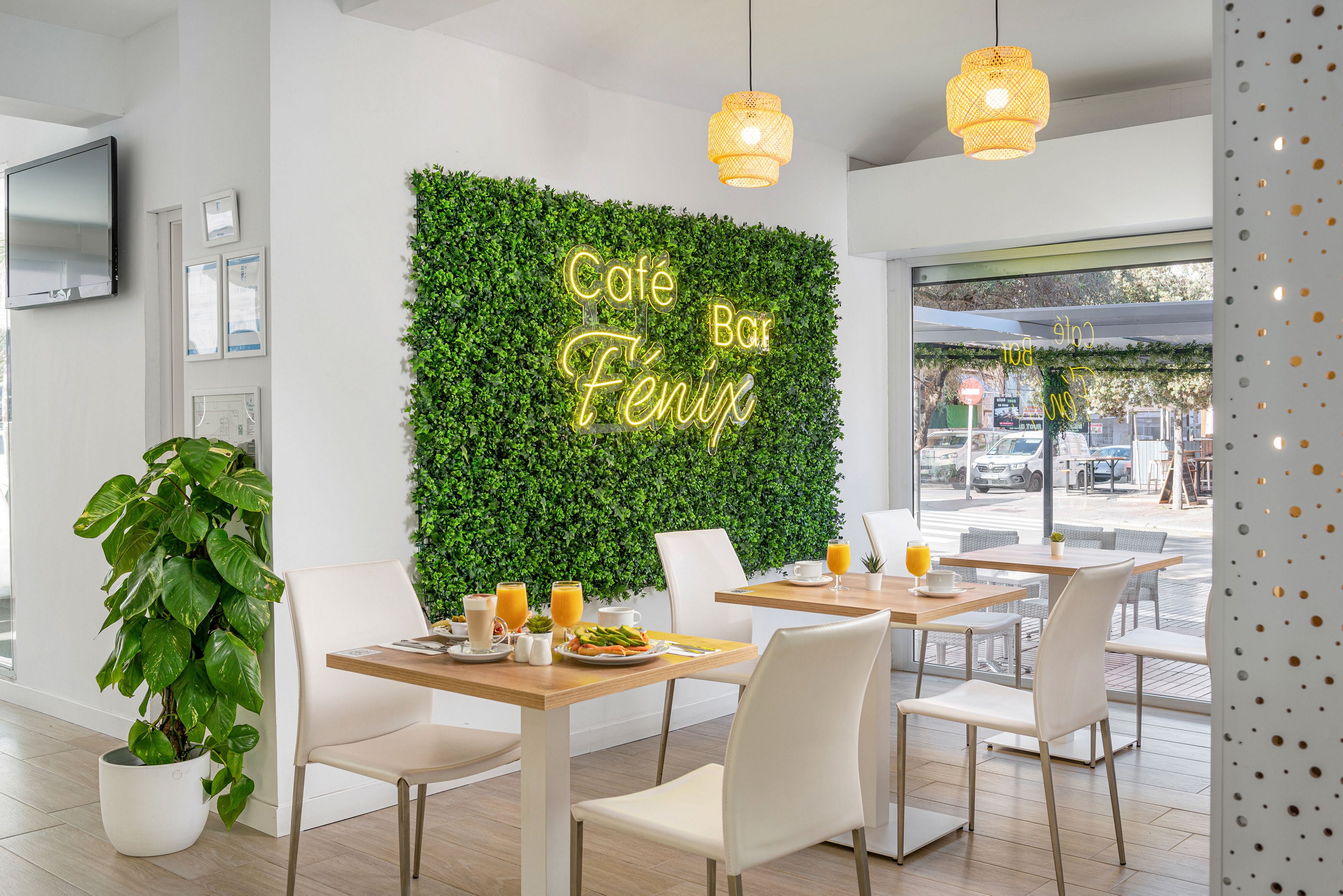 Contemporary café interior featuring a green plant wall with neon text reading Café Bar Fenix. Light wooden tables are set with glasses of orange juice and breakfast dishes, surrounded by white chairs. Large windows let in natural daylight and reveal an outdoor street view. Warm pendant lights and indoor plants create a fresh, inviting atmosphere.