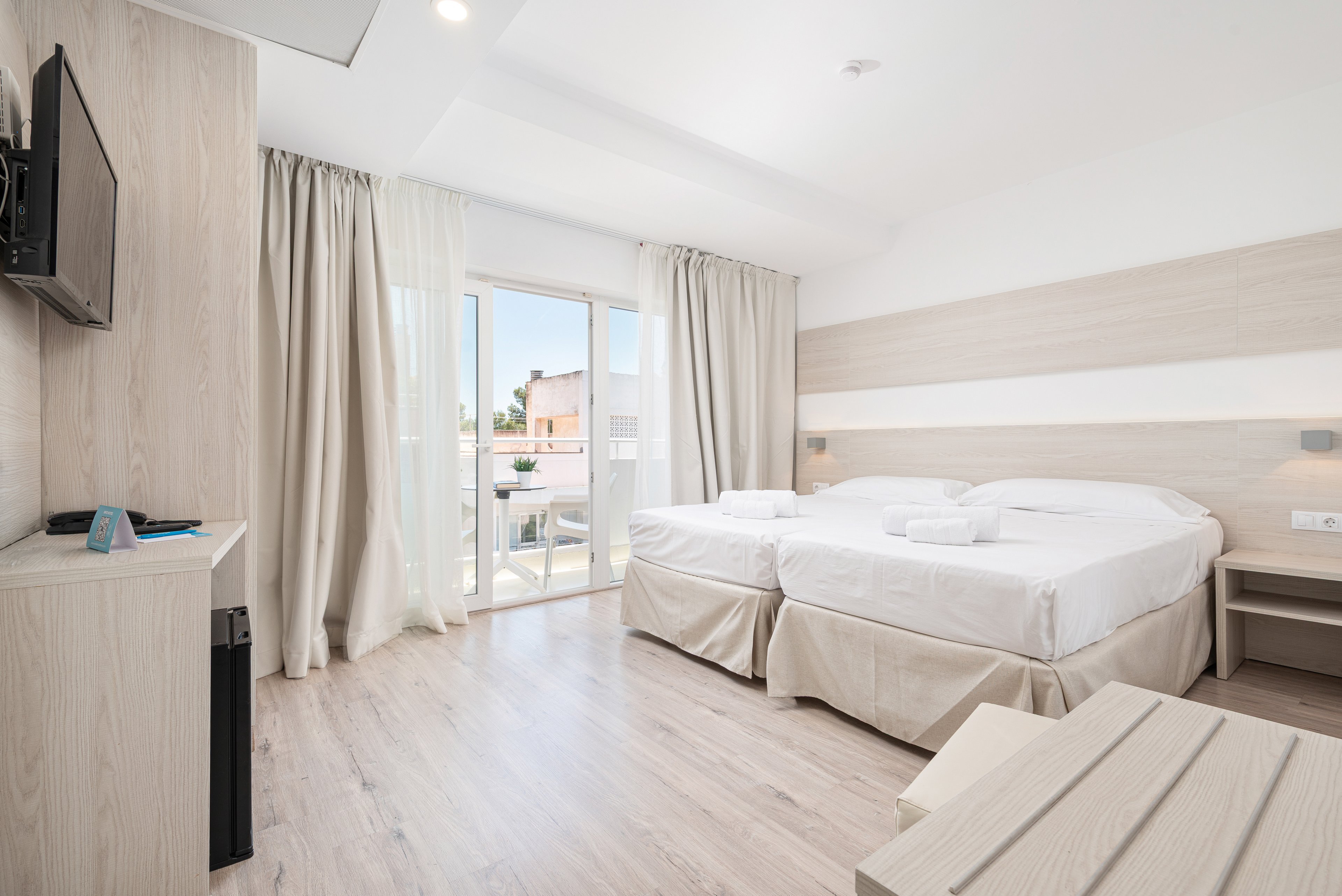 A bright, modern hotel room features two neatly made twin beds with white linens and folded towels on top. Light wood furniture and flooring create a clean, minimalist aesthetic, complemented by neutral curtains and walls. A flat-screen TV is mounted on the wall opposite the beds, and a small desk area sits nearby. Large glass doors open onto a balcony, allowing natural light to fill the space and offering a view of neighboring buildings.