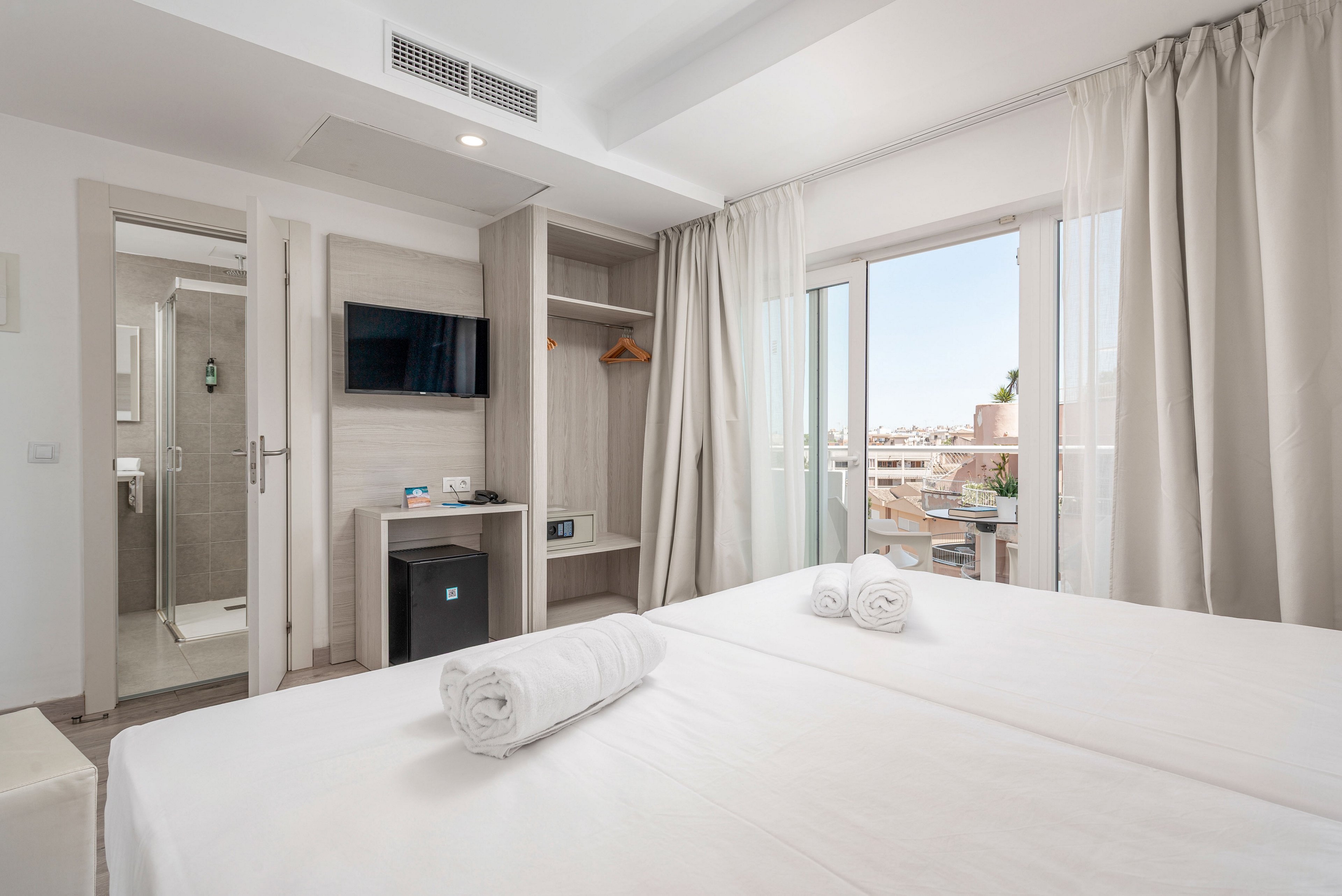 A bright, modern hotel room features a neatly made double bed with white linens and rolled towels. The space includes a wall-mounted flat-screen TV, a small desk area, and a compact refrigerator beneath. An open door reveals a clean, tiled bathroom, while large glass doors lead to a balcony overlooking city buildings. Soft neutral tones and natural daylight create a calm, inviting atmosphere.
