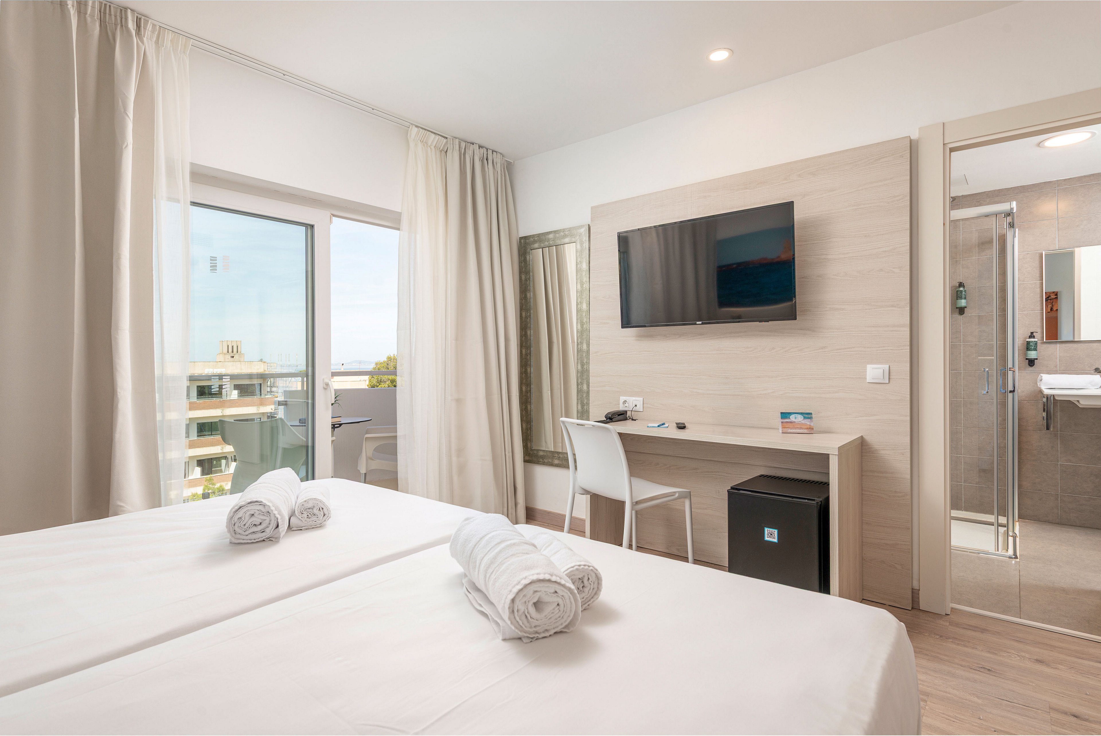 A clean, modern hotel room features two neatly made twin beds with rolled white towels placed on top. A desk with a chair faces a wall-mounted flat-screen TV, with a small refrigerator positioned underneath. Floor-to-ceiling curtains frame a glass door leading to a balcony overlooking nearby buildings and greenery. To the right, a glass door opens into a contemporary bathroom, completing the airy and minimalist interior design.