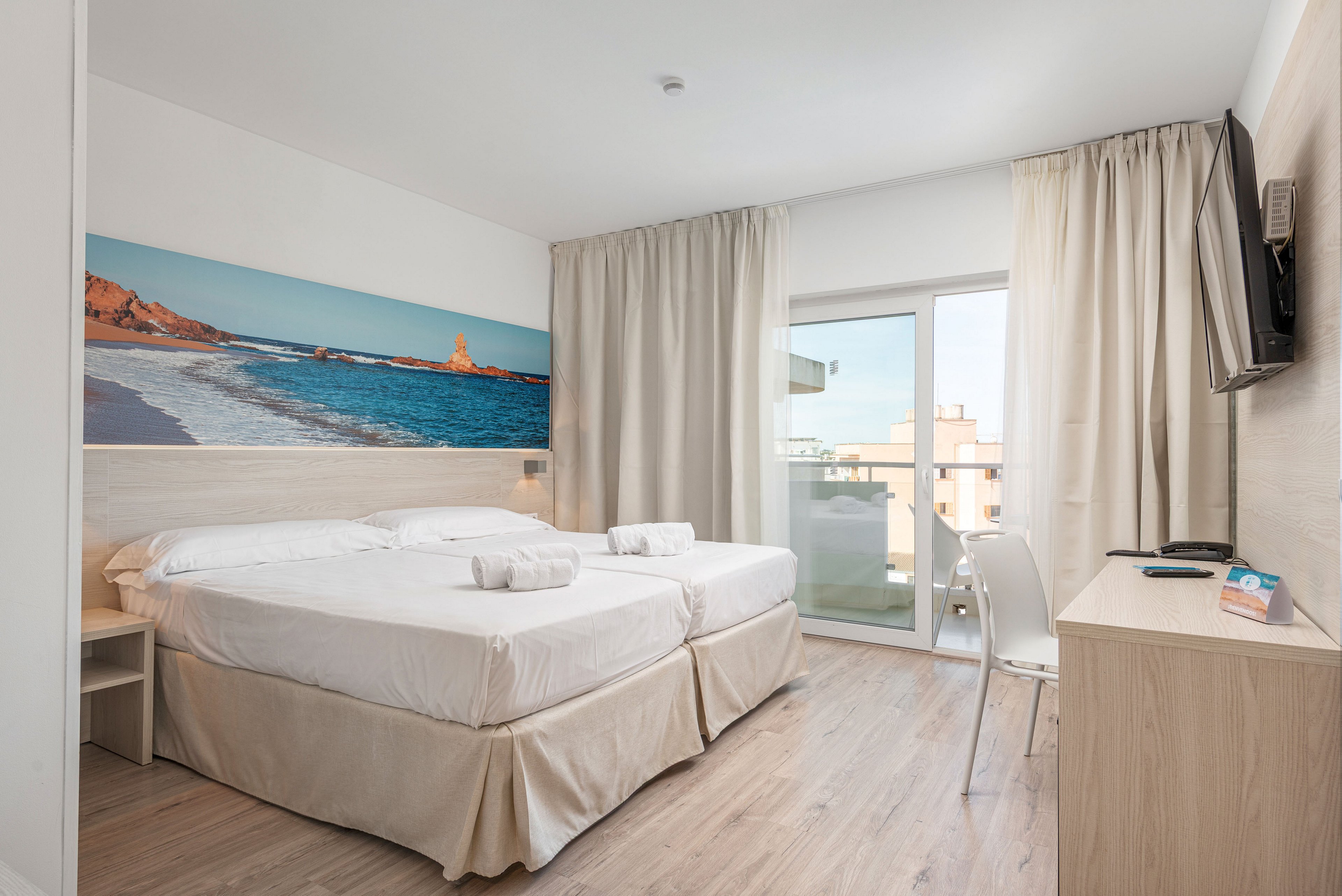 A clean, modern hotel room features two neatly made beds with white linens and folded towels. Light wood flooring and furniture complement the neutral walls and curtains, creating an airy atmosphere. A large beach landscape mural adds color above the beds, while a flat-screen TV is mounted on the wall opposite. Floor-to-ceiling glass doors open to a small balcony with views of nearby buildings, letting in natural daylight.