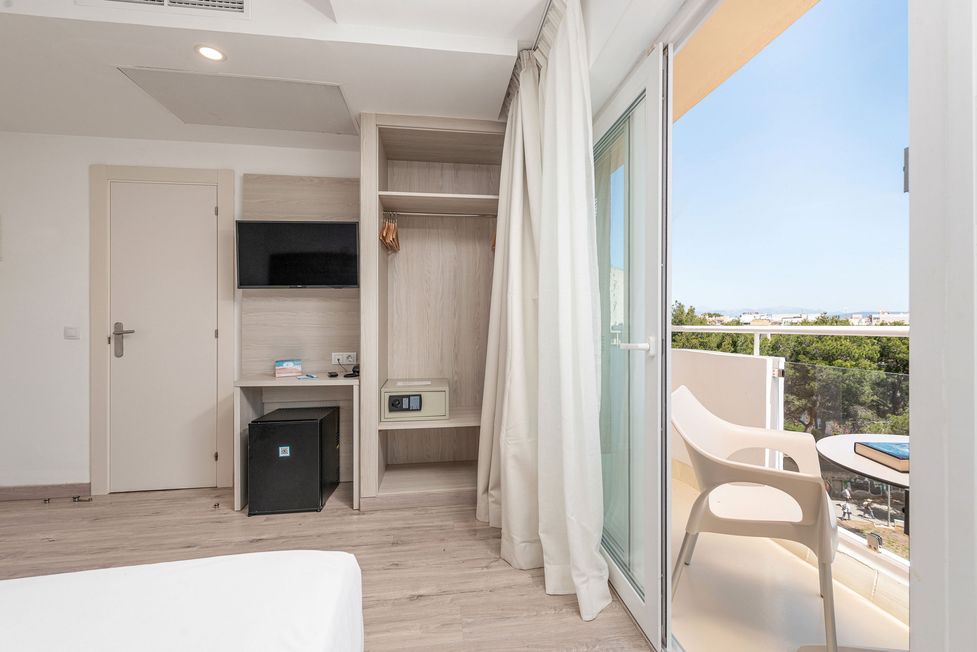 Modern hotel room interior featuring a wall-mounted television, open wardrobe, and small desk with a mini fridge and safe. Light wood flooring and neutral walls create a clean, airy atmosphere. A large glass door opens to a balcony with two contemporary chairs and a table overlooking trees and distant buildings. Daylight floods the space through the balcony, emphasizing the minimalist, vacation-ready design.