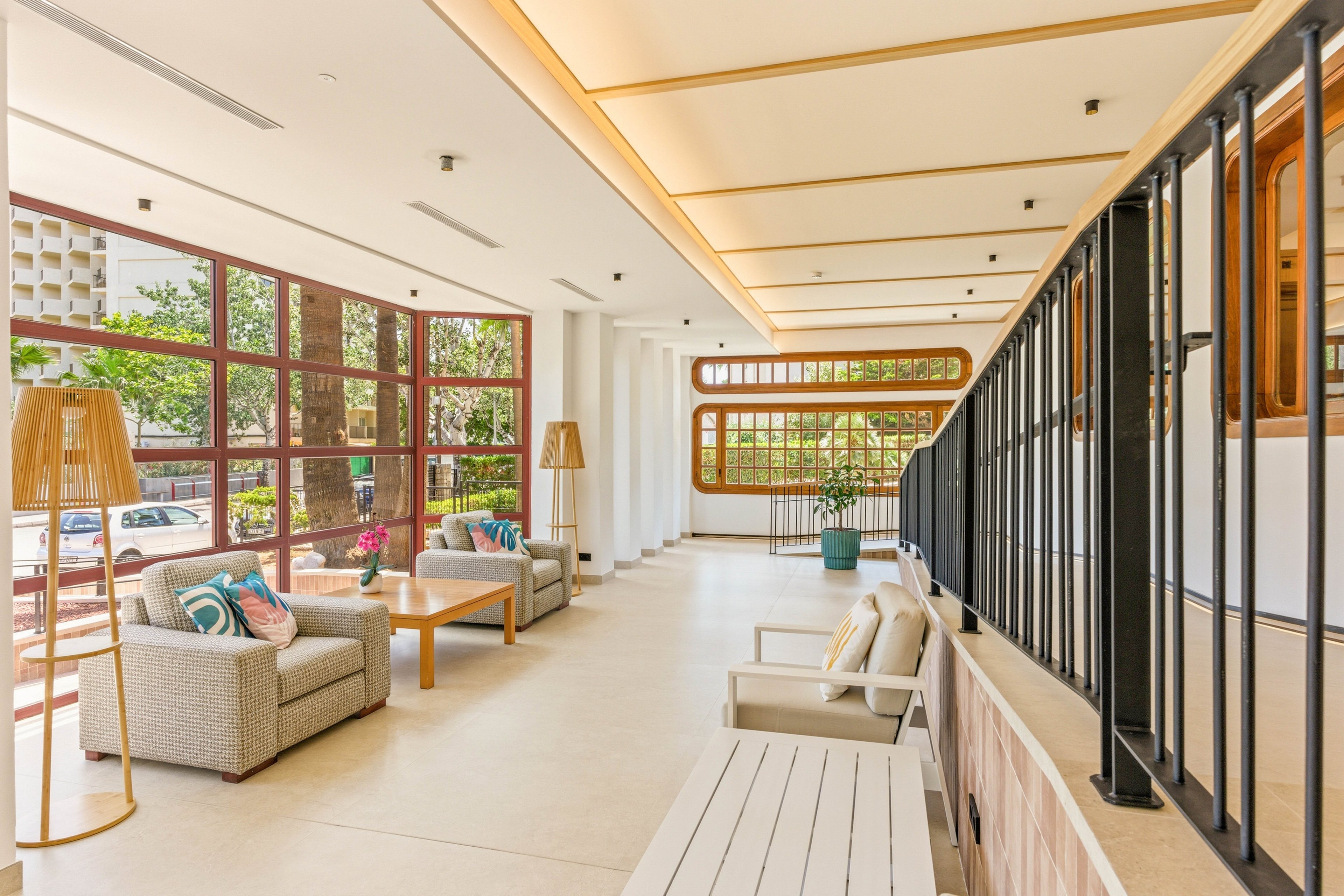 A spacious modern lounge area features comfortable armchairs and sofas arranged around wooden coffee tables. Floor-to-ceiling windows line one side, allowing abundant natural light and views of greenery and parked cars outside. Neutral tones dominate the interior, accented by colorful cushions, flowers, and tall floor lamps. The clean, minimalist design and open layout create an inviting, contemporary social or waiting space.