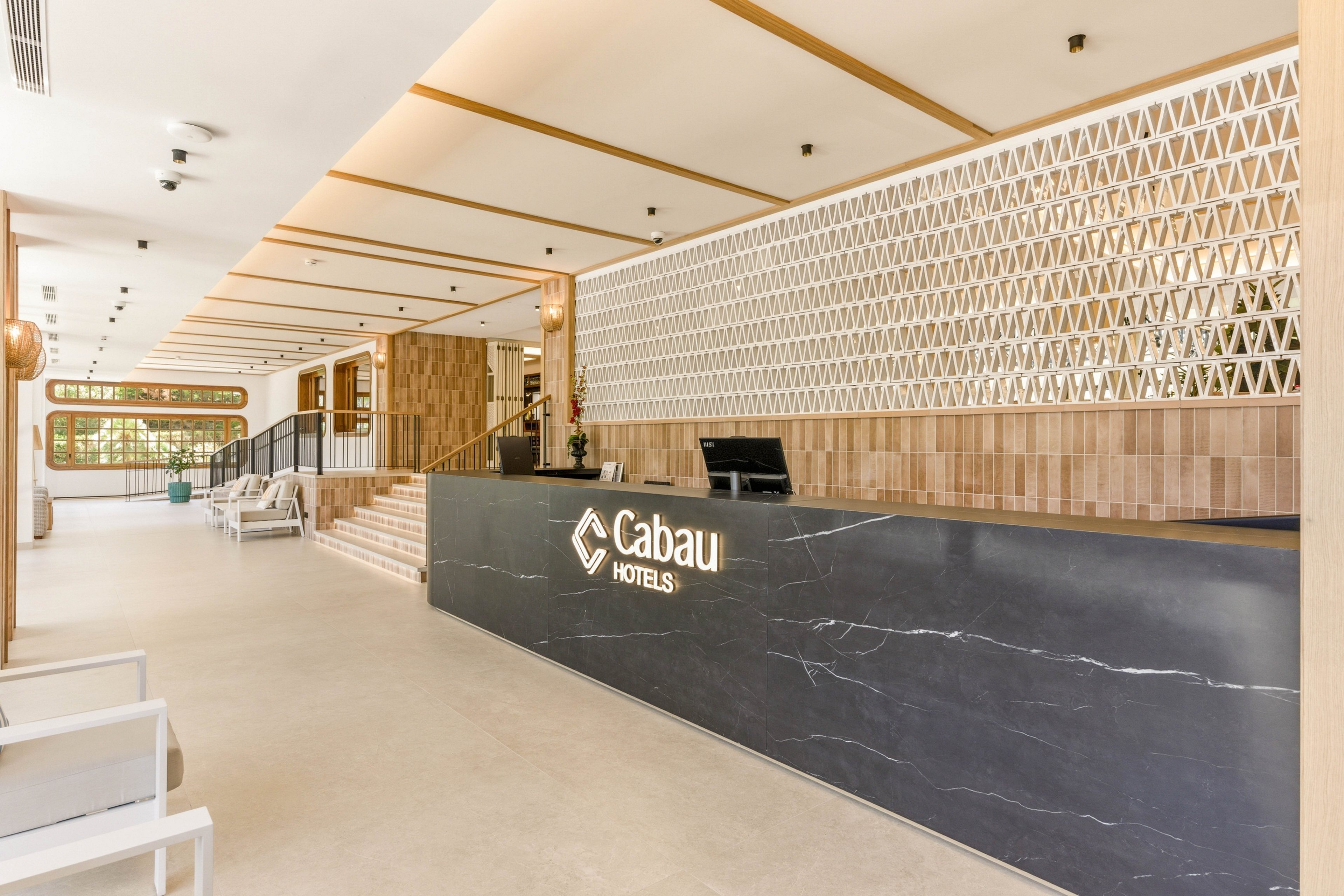Spacious hotel reception area featuring a long black marble front desk with illuminated Cabau Hotels text. The lobby has light wood paneling, geometric wall patterns, and a minimalist seating area. Stairs lead to an upper level, and large windows bring in natural light. The overall design feels contemporary, clean, and welcoming.