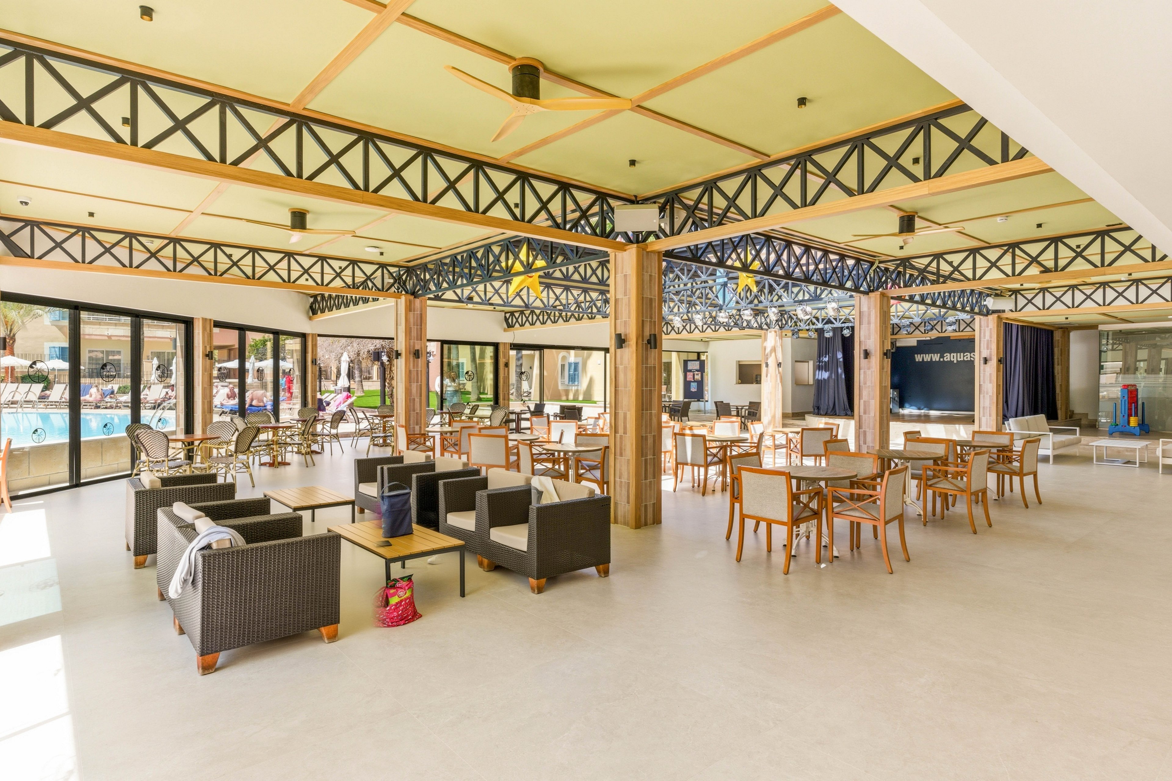 Bright, open resort lounge area with modern wicker and wooden seating is arranged across a large tiled floor. Floor-to-ceiling windows and glass doors reveal an outdoor swimming pool and sunbathers in the background. Exposed beams and wooden columns add a contemporary, airy feel to the interior. The space appears ready for guests to relax, socialize, or enjoy refreshments near the pool.
